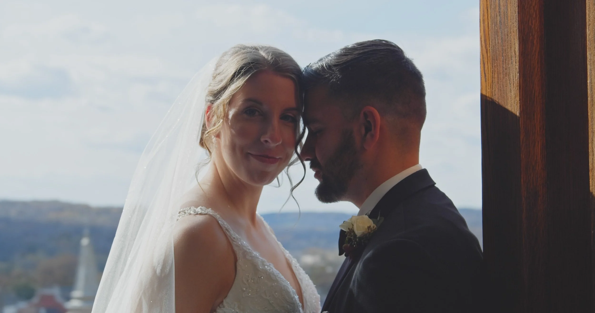 Bride and groom share a moment during their wedding, with the bride wearing a veil and wedding dress, and the groom in a suit, standing close together indoors near a wooden wall with a scenic view in the background.