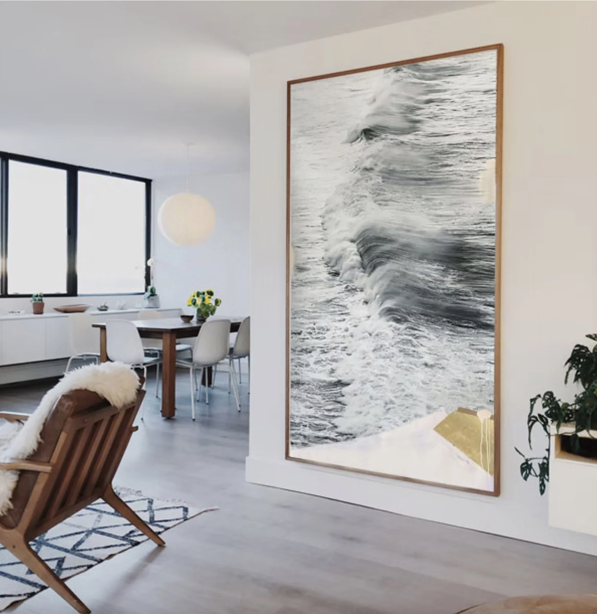 Living room with a large wall art photo of ocean waves, dining table with chairs, wooden lounge chair with a sheepskin throw, and large windows with a view of the outside.