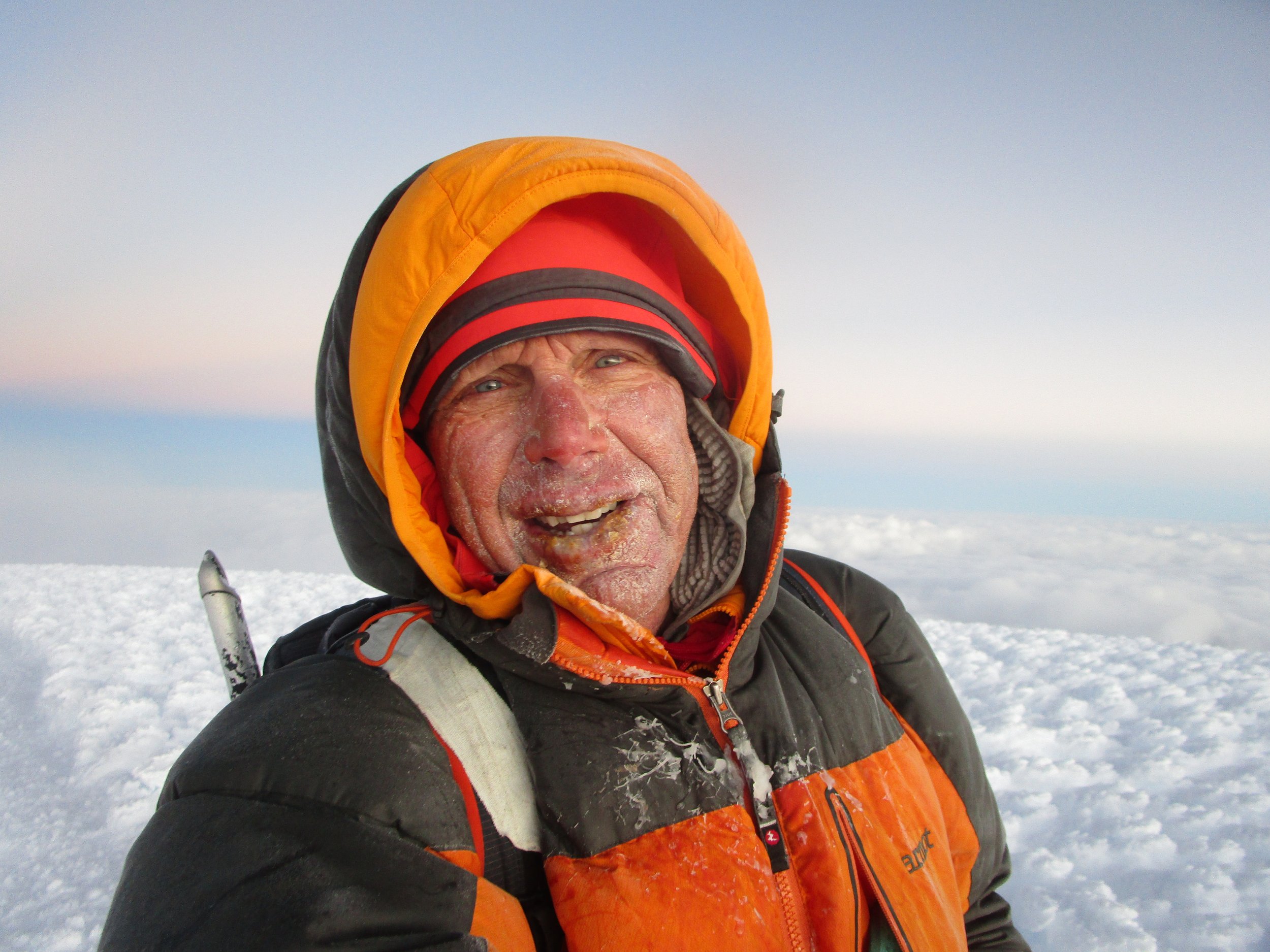 A man in winter gear atop a snow-covered mountain, with ice and snow on his face, wearing a black and orange jacket and a red hat, against a sky with clouds.