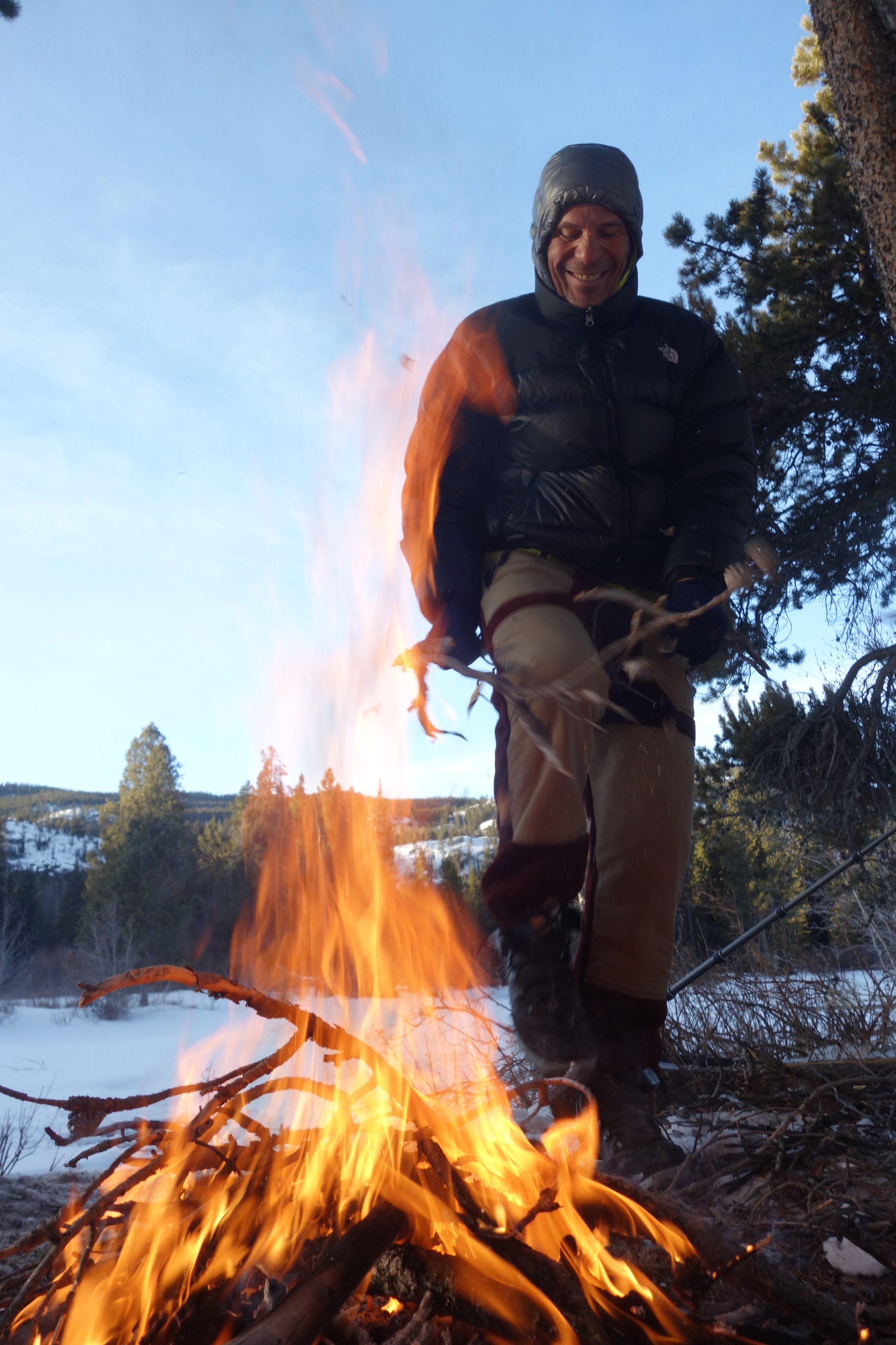 A person dressed in outdoor winter clothing warming himself near a campfire in a snowy forest landscape.