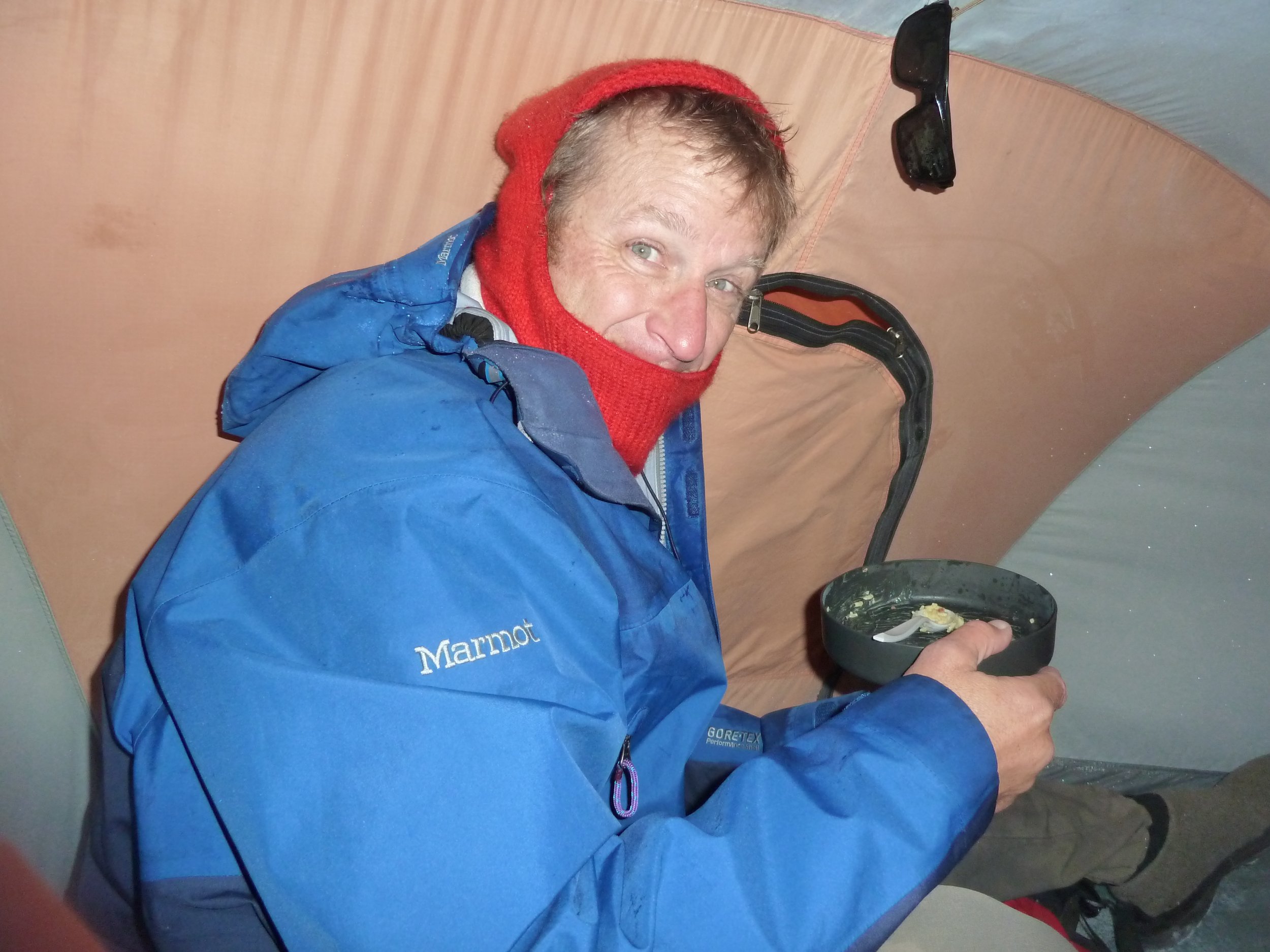 A person with light-colored hair, blue eyes, wearing a red knit hat and blue jacket, sitting inside a tent and eating from a black bowl.