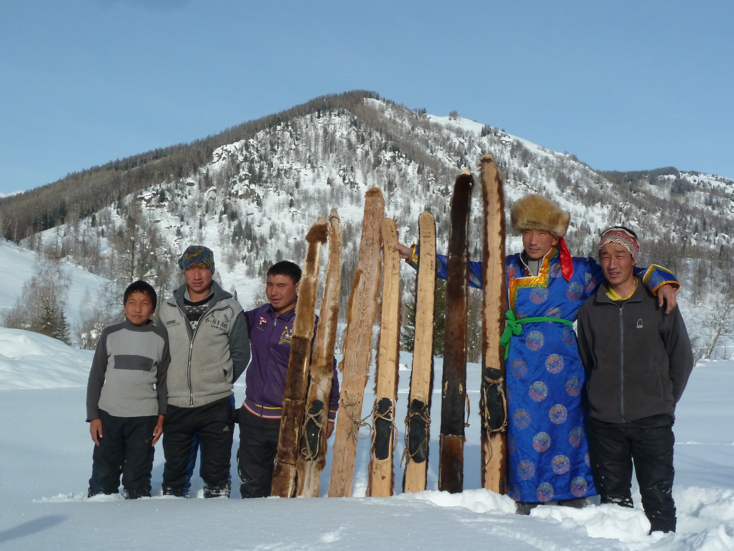 A group of six people standing in snow with a mountain and trees in the background, holding or standing near traditional wooden skis, with one person dressed in colorful traditional clothing.