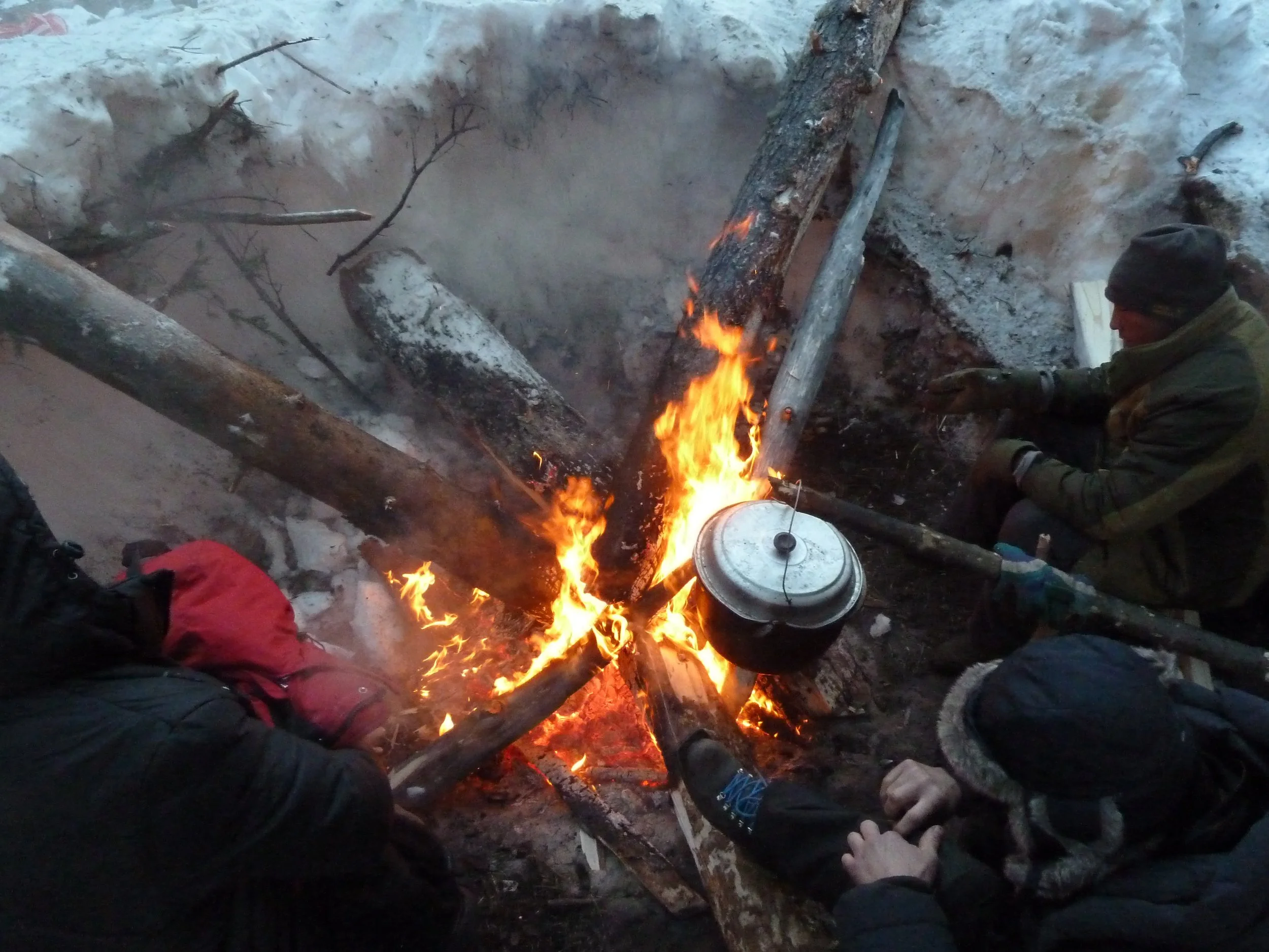 People gathered around a campfire, cooking food in a pot outdoors in a snowy environment.