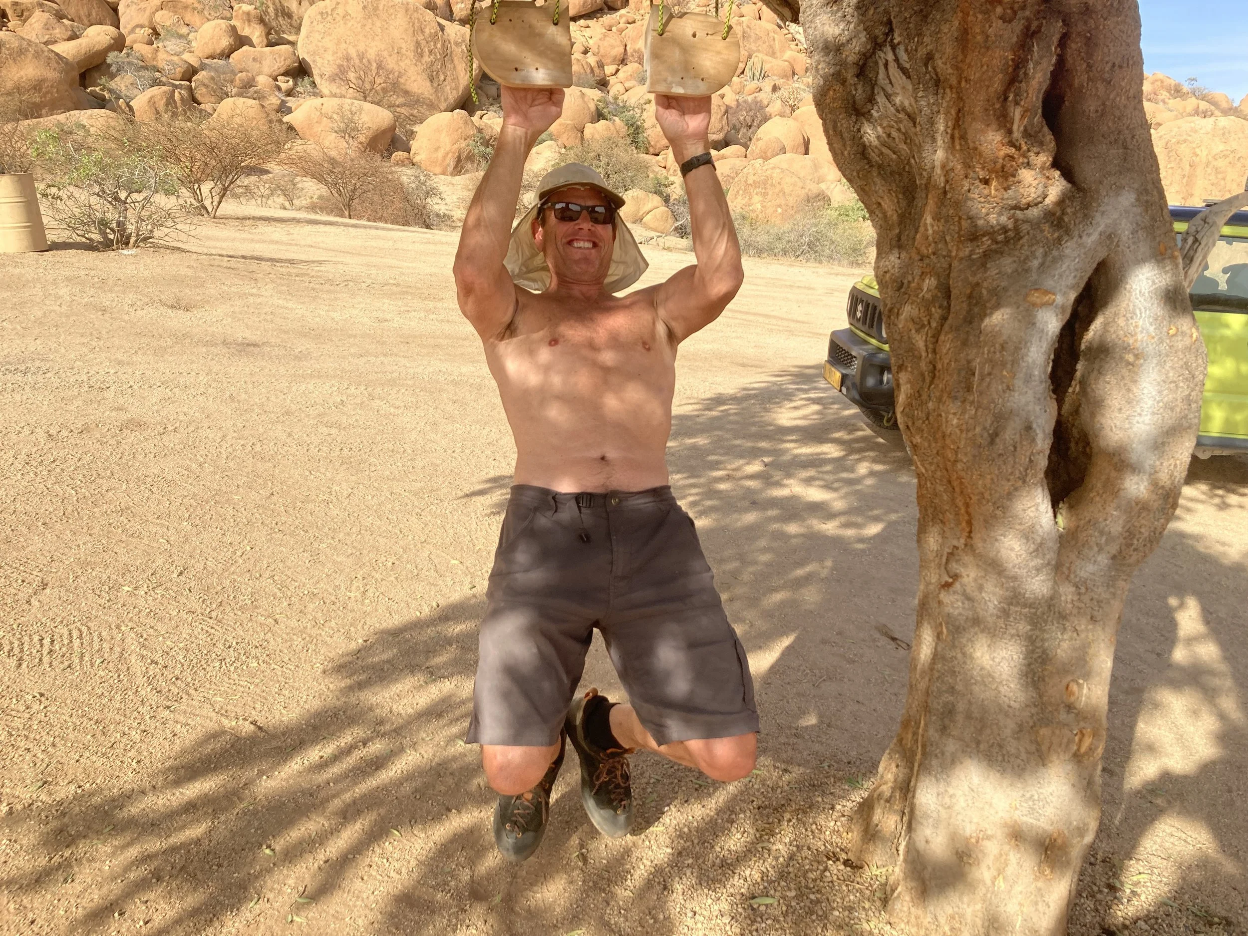 A shirtless man wearing sunglasses, a hat, and shorts is hanging from a tree branch in a desert landscape with rocks and sparse vegetation.
