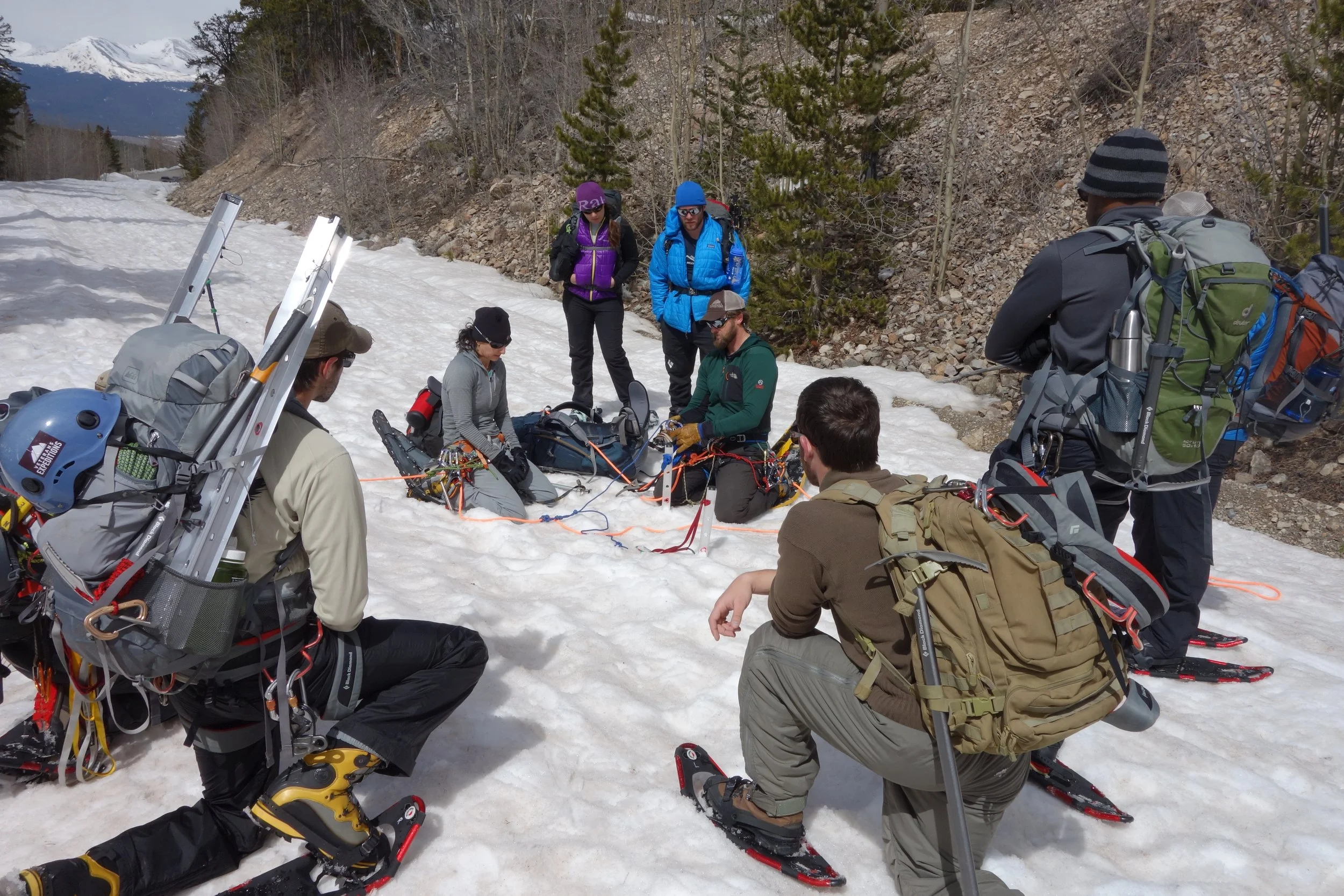 Group of climbers on snow-covered terrain, surrounded by trees and mountains, preparing for mountaineering or rescue activity.