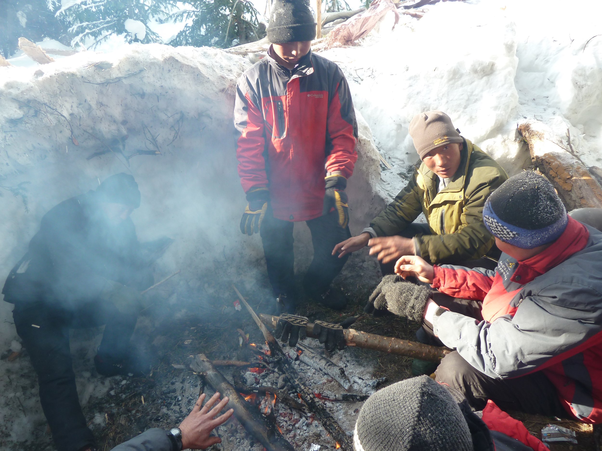 Group of four people outdoors in snowy environment gathered around a small campfire, some tending to the fire and others observing.