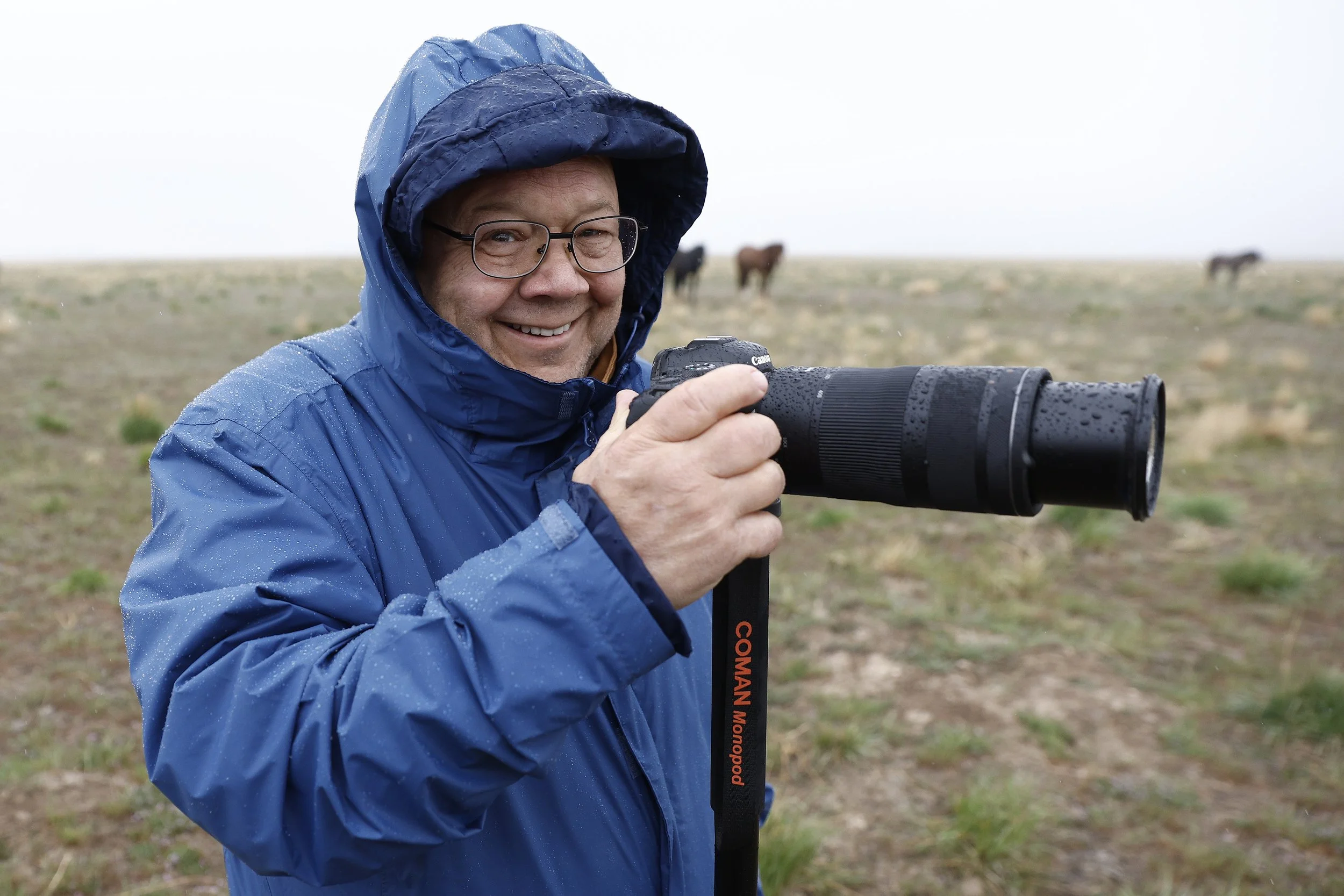 Man in a blue rain jacket with a hood, holding a camera with a long lens, standing outdoors in a grassy field with horses in the background on a rainy day during a wild horse photography workshop.