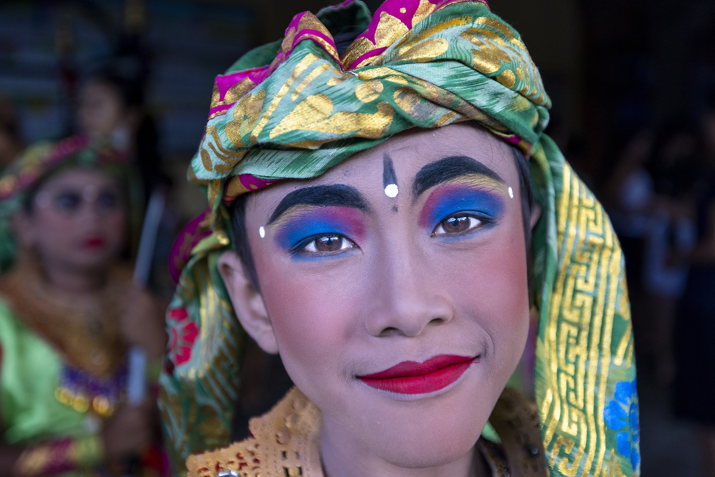A person in traditional colorful attire with ornate makeup, including bright eyeshadow, bold eyebrows, and red lipstick, smiling directly at the camera. They are wearing a patterned headdress and other traditional clothing in Bali Indonesia.