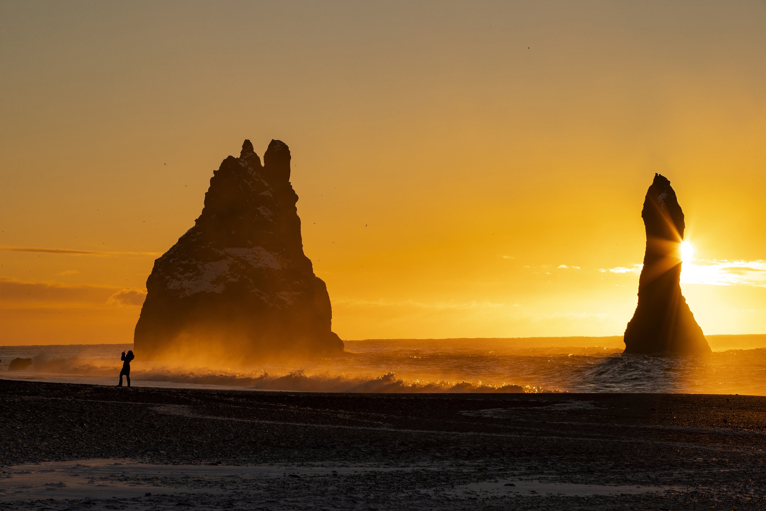 Silhouette of sea stacks at sunset with a person taking photos on black beach in Iceland.