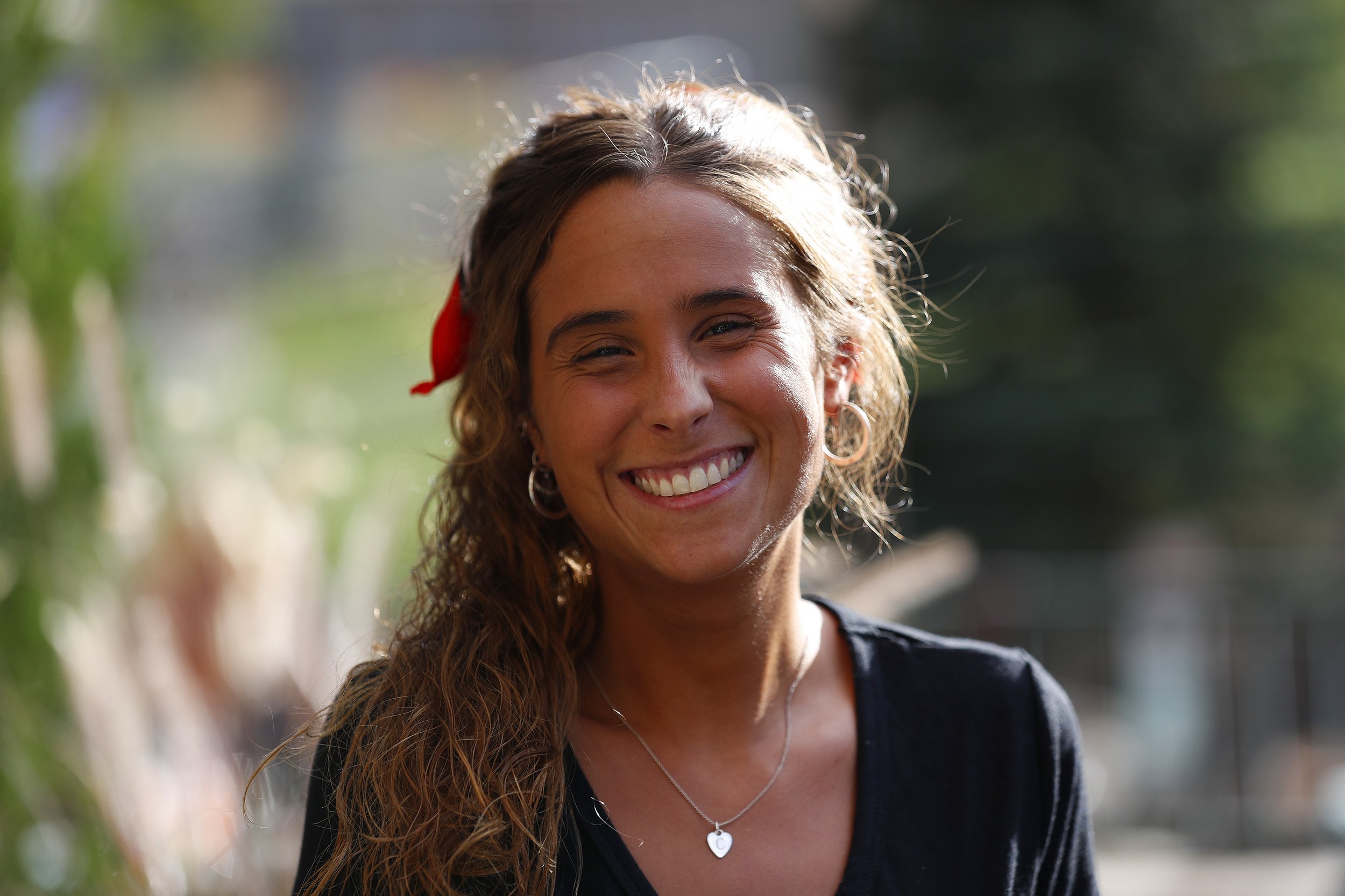 Smiling woman with wavy hair and a red ribbon, wearing hoop earrings and a heart-shaped necklace, in an outdoor setting.