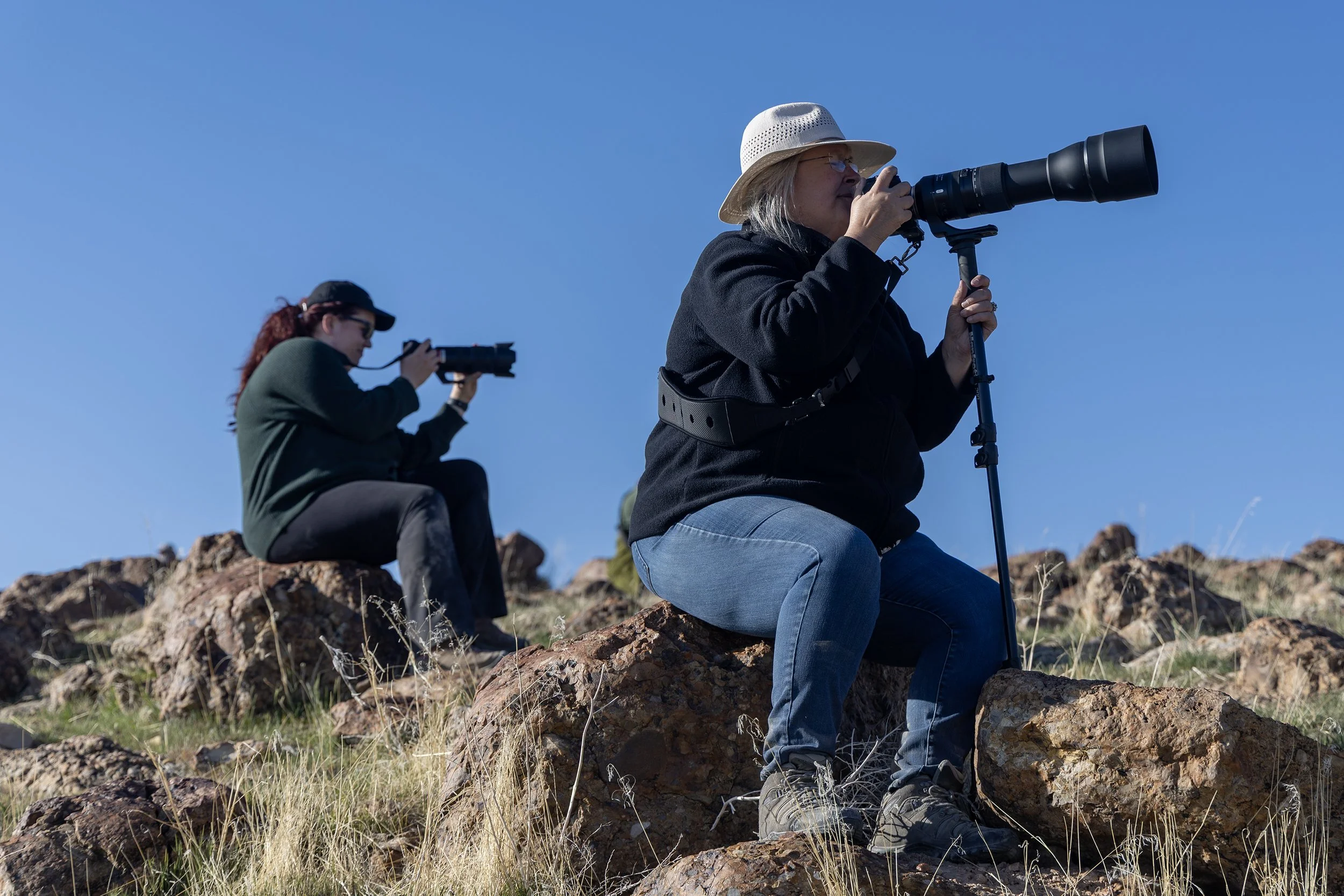Two women sitting on rocks outdoors, using binoculars and a camera with a long lens, under a clear blue sky.