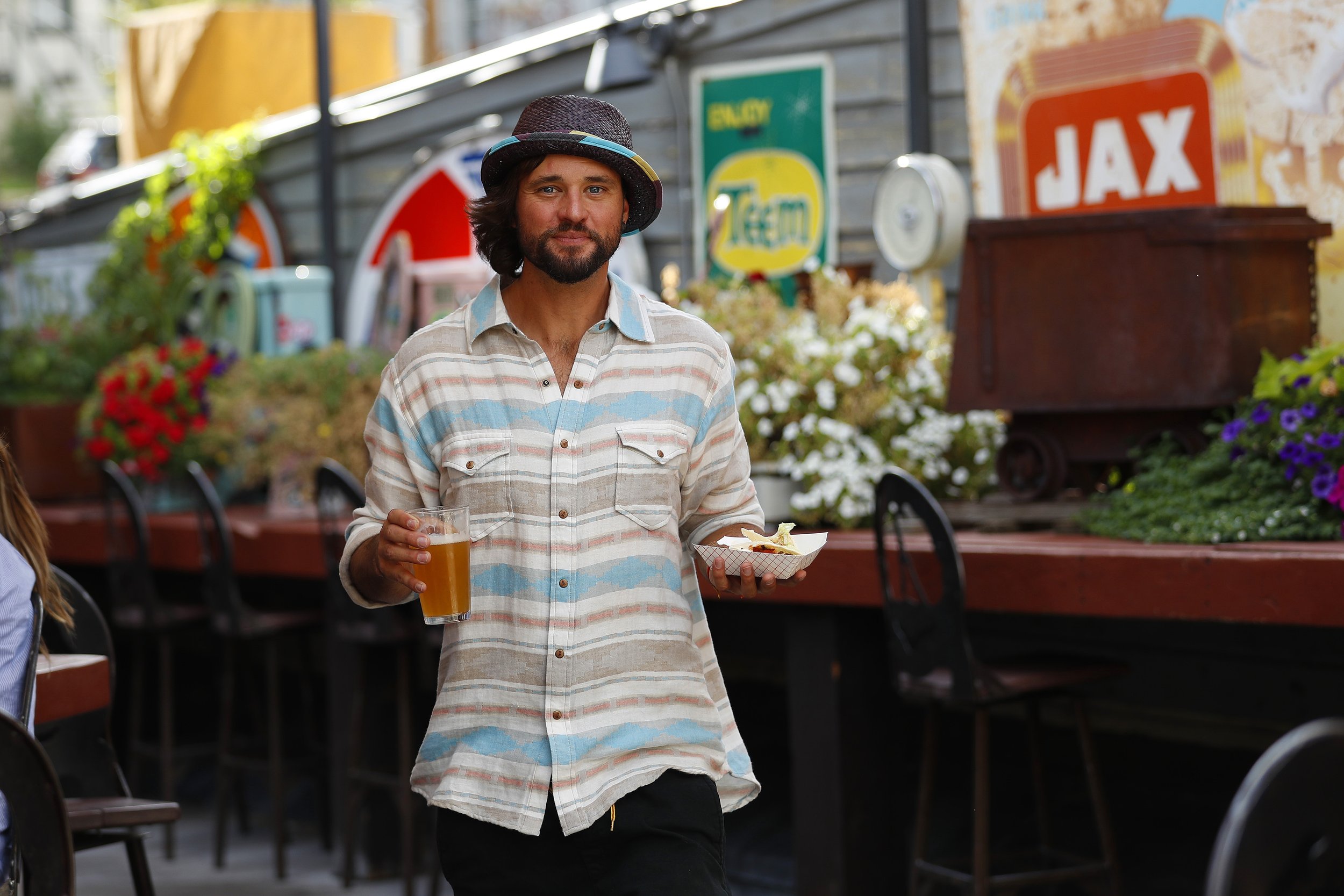 Man with a hat holding a beer and food at an outdoor eatery