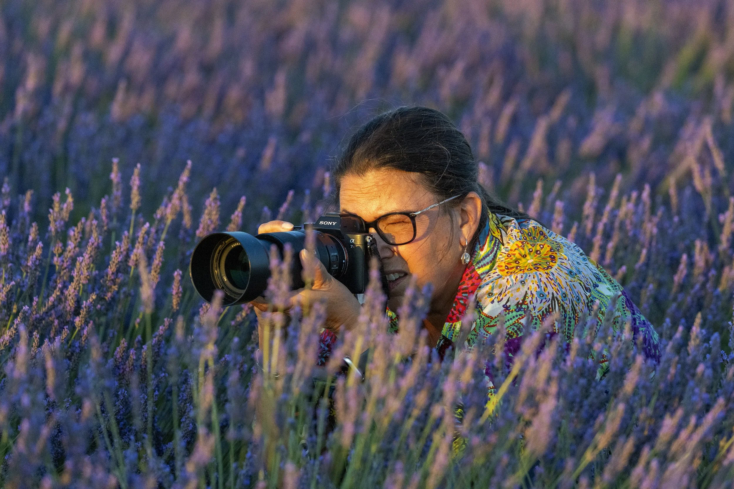 A woman wearing glasses and a colorful shirt is taking a photograph with a camera in a lavender field during sunset.