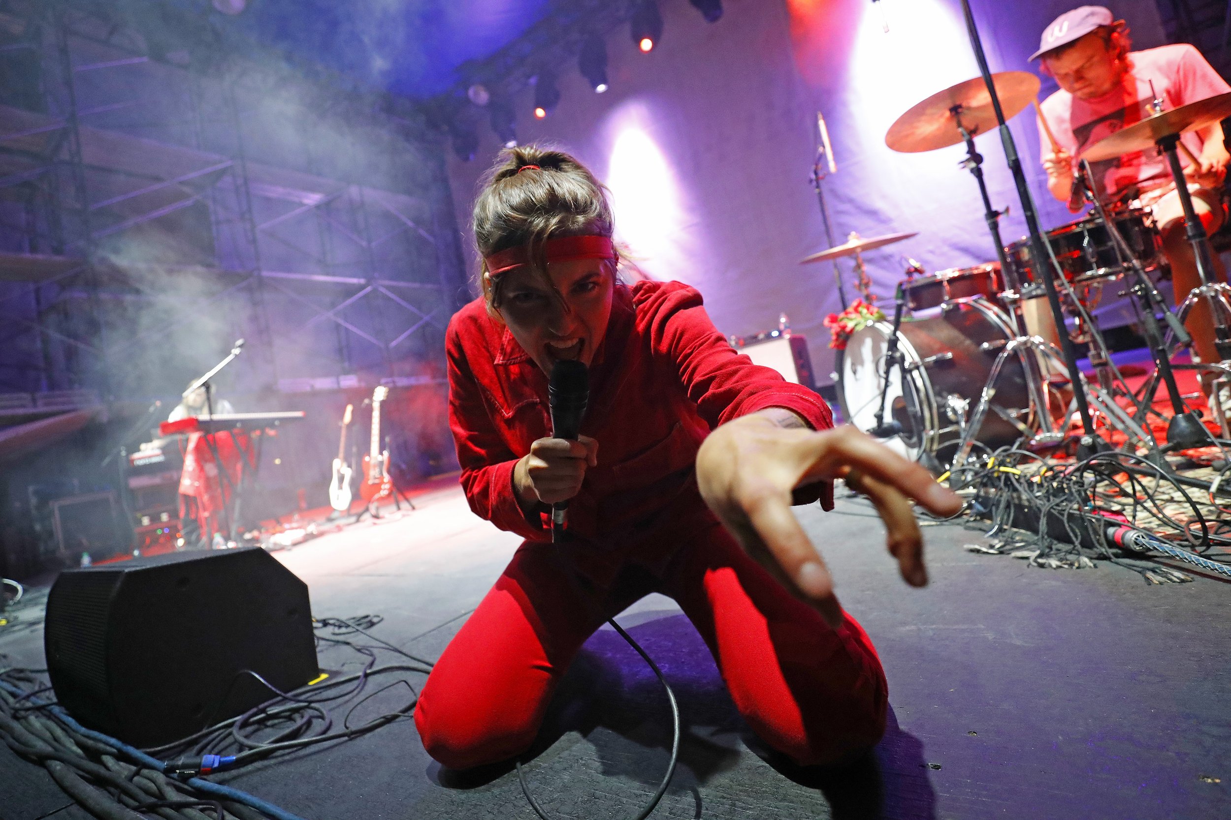A performer kneeling on stage with a microphone, reaching towards the camera in a dynamic pose. The stage is equipped with musical instruments and a band member is visible playing drums durig my event coverage of the Utah Arts Festival.