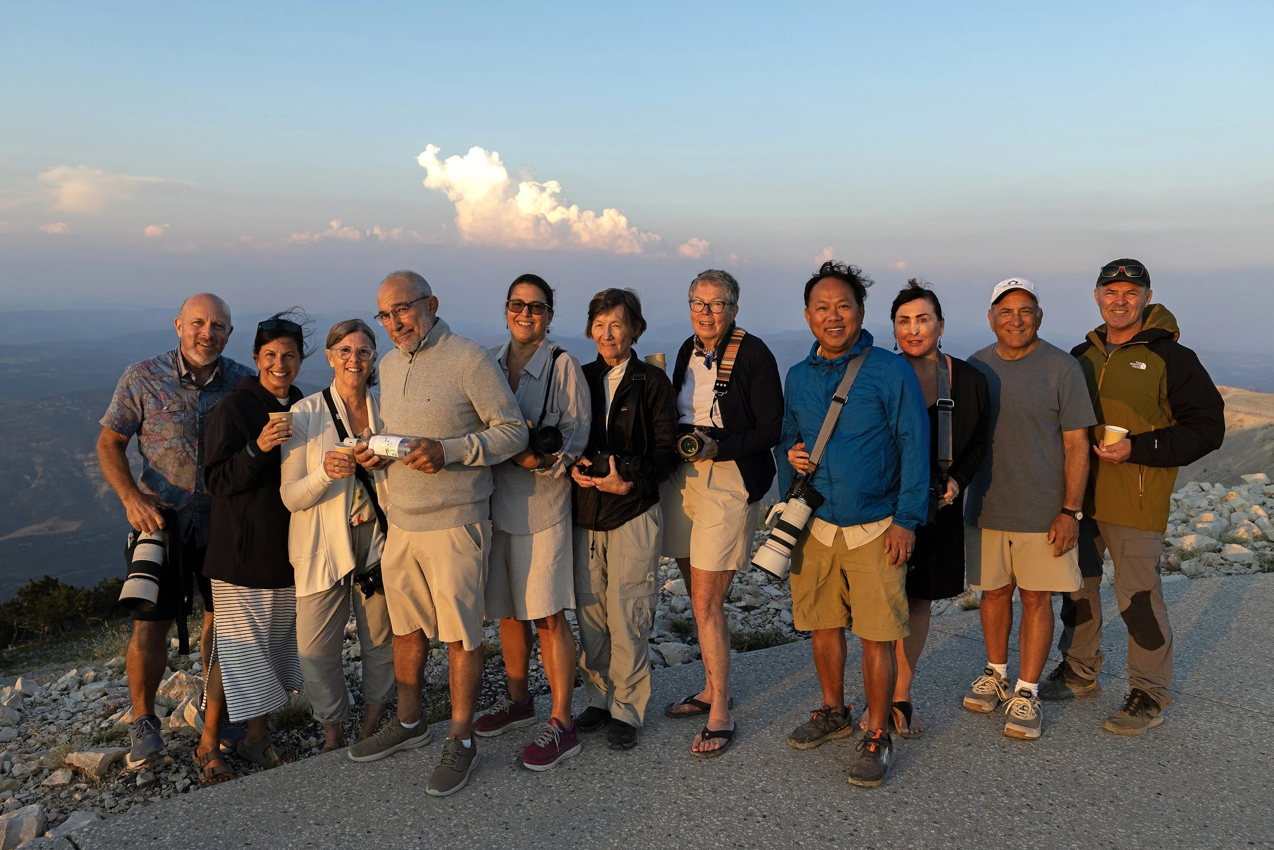 Group photo of photography workshop participants on the summit of Mont Ventoux in Provence, France at sunset, smiling and holding cameras and drinks during a photo adventure.