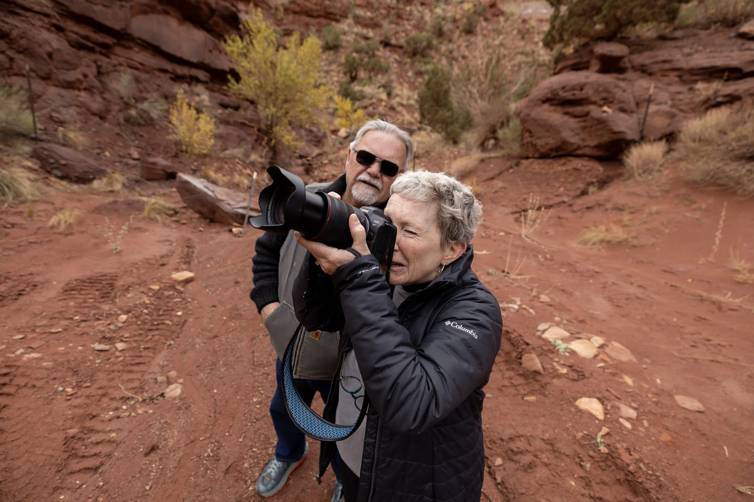 An elderly woman takes a photo with a professional camera while an elderly man stands behind her on a rocky, desert landscape with sparse bushes and yellowing trees in Moab.