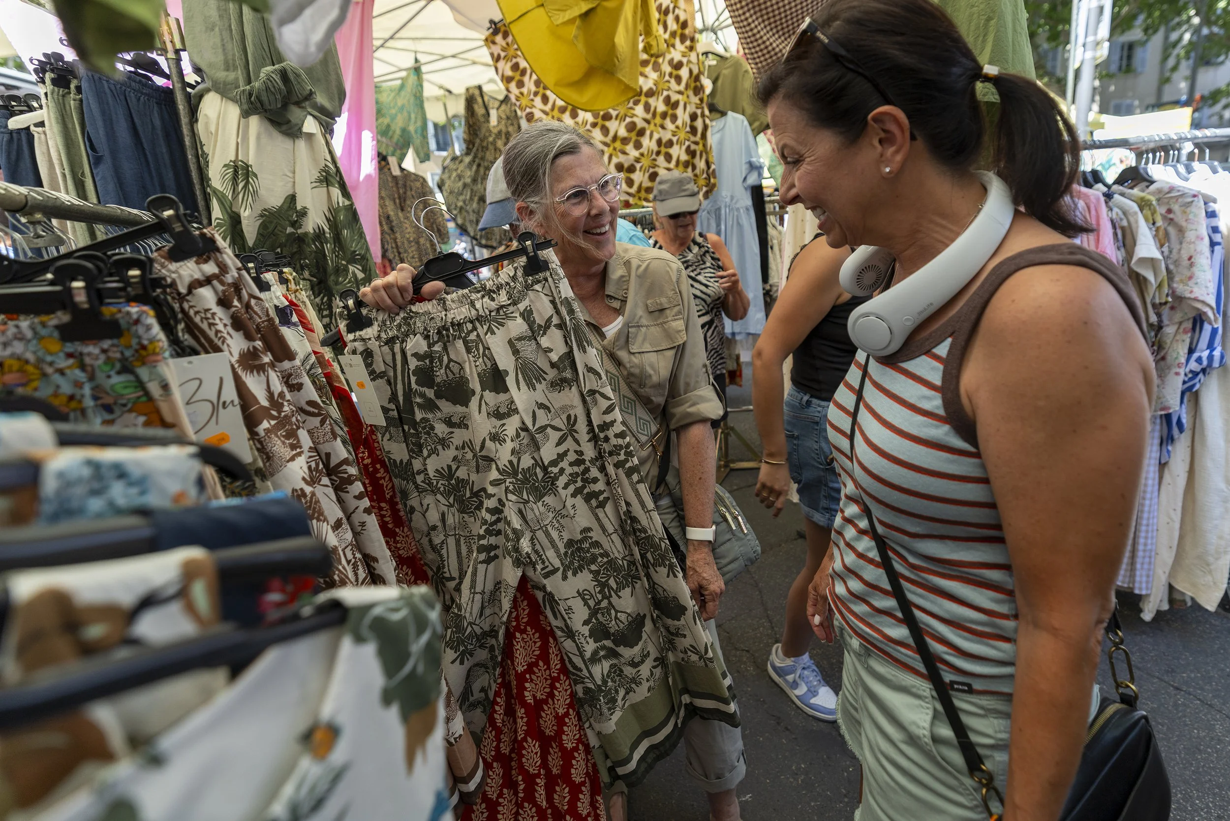 Two women are shopping at an outdoor clothing market, smiling and laughing while looking at floral skirts. One woman is holding a skirt up for the other to see, with clothing racks and other shoppers in the background. in France.