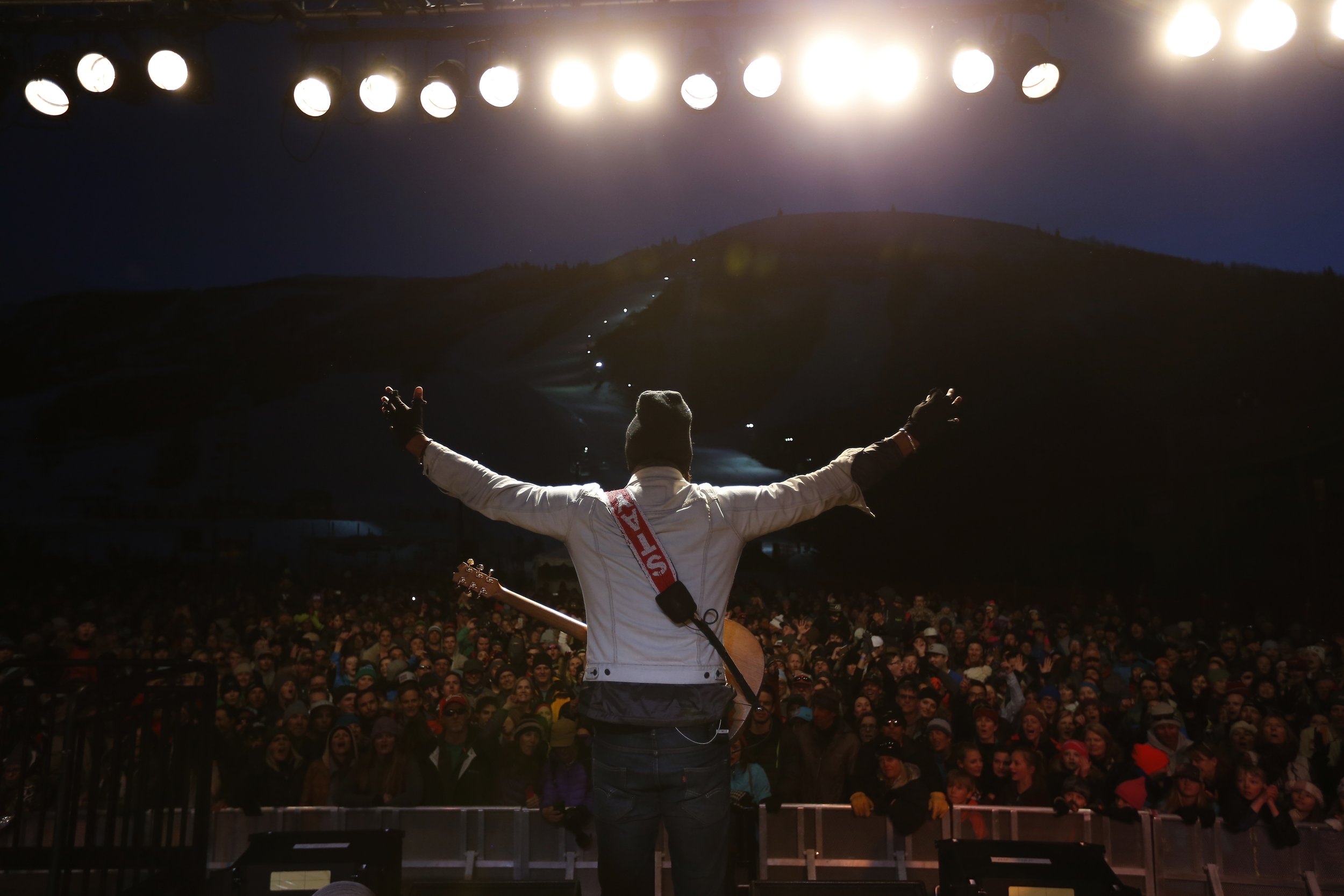 Musician Michael Franti performing on stage at night in front of a large crowd, with bright stage lights and a mountain in the background.