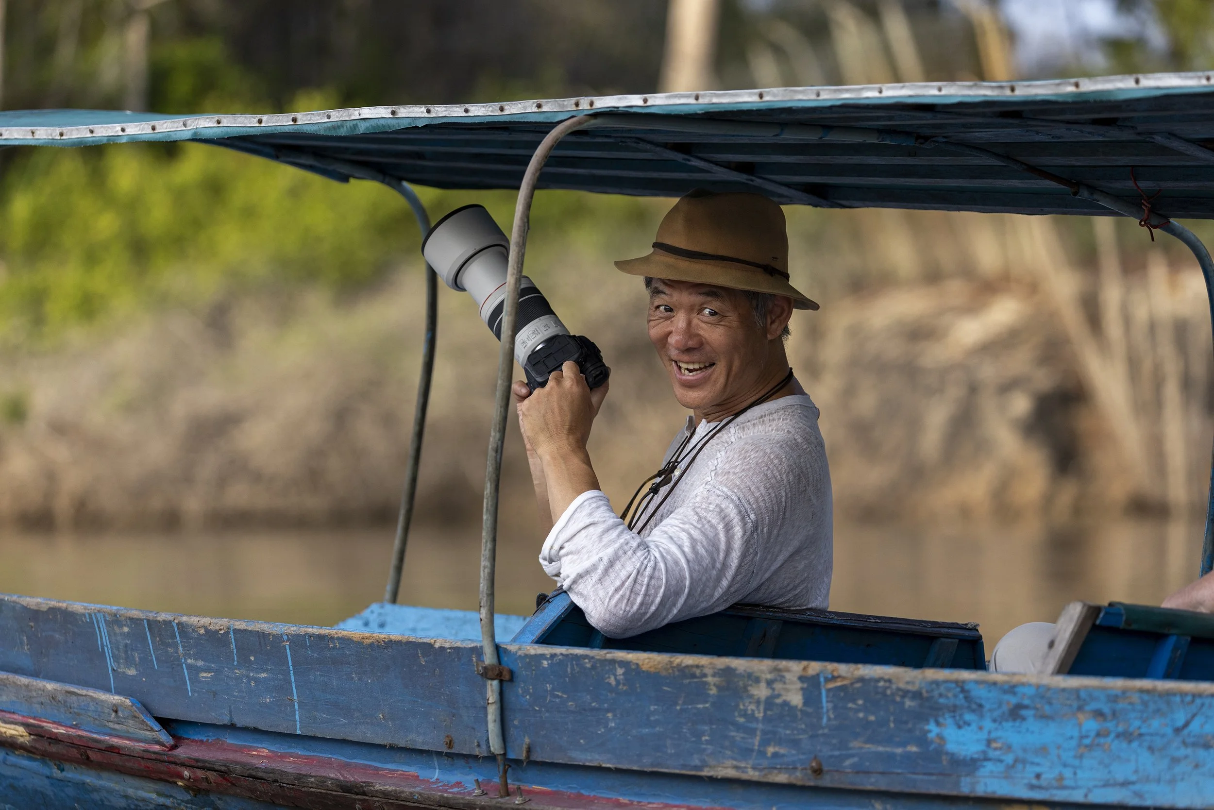 Photography workshop participant taking photos from a long boat in Borneo, Indonesia, capturing the lush rainforest and river scenery during a guided photo tour.