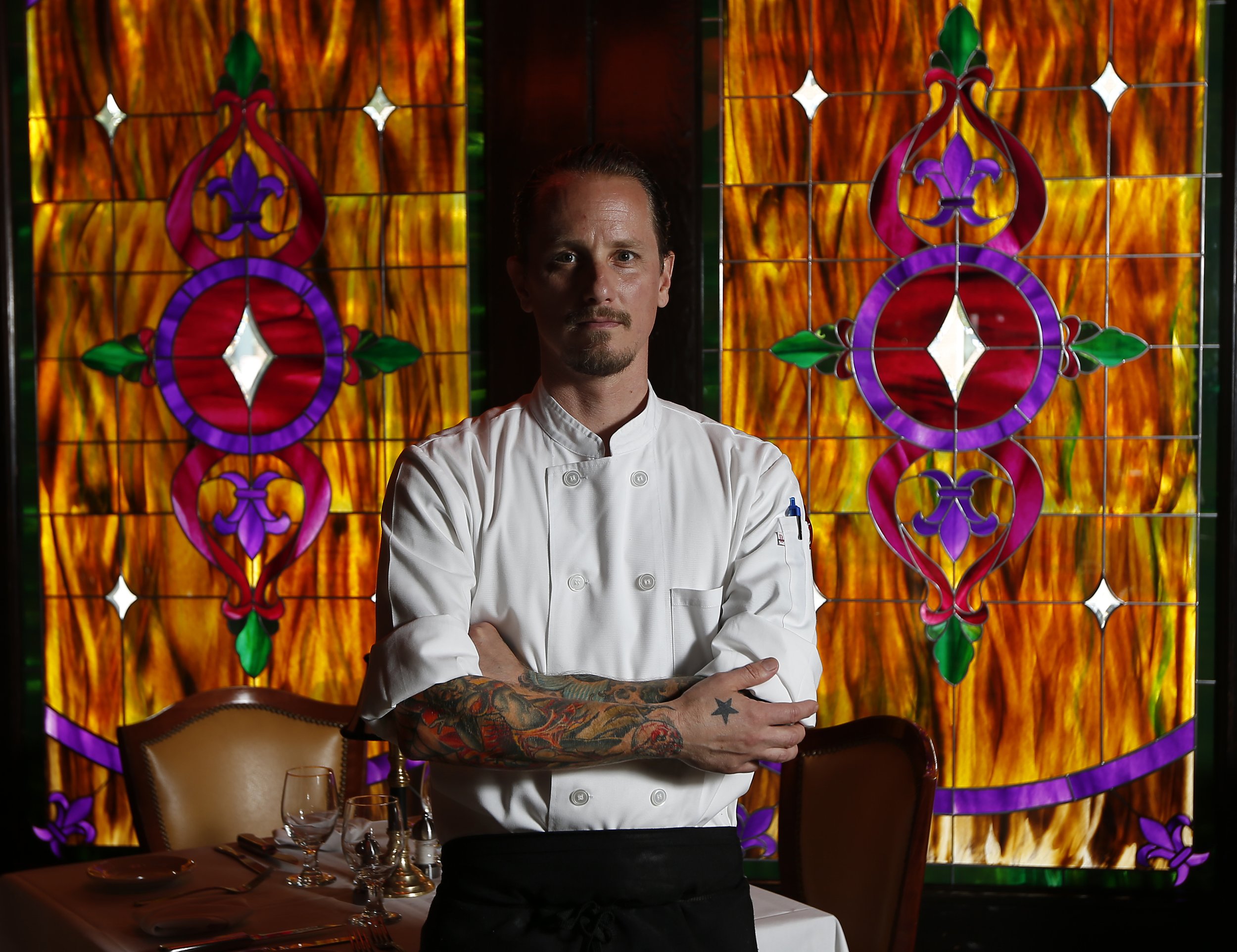 Chef with tattooed arms standing in front of a stained glass window in a restaurant for a restaurant review.