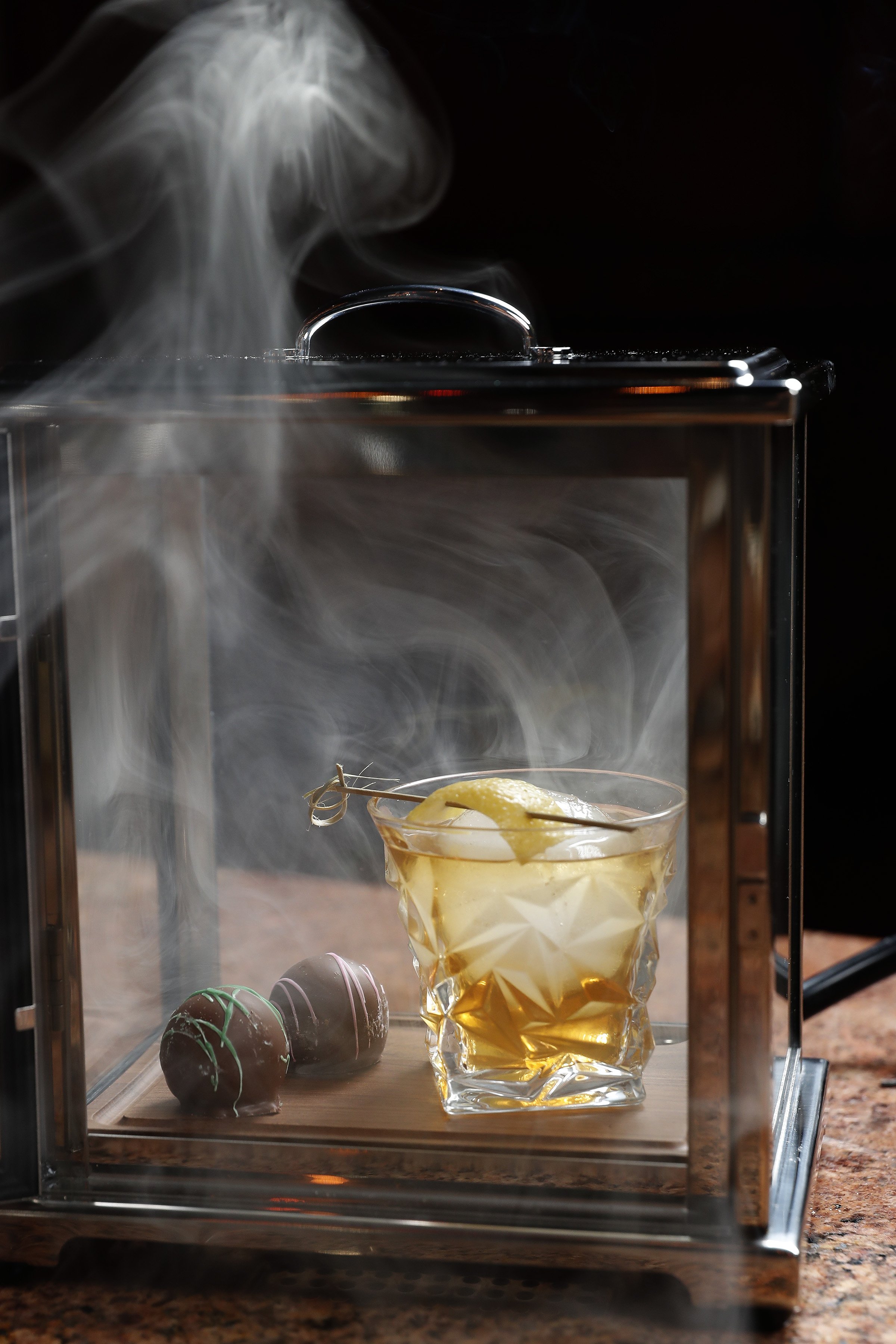 A smoked cocktail with garnished lemon peel in a glass, placed inside a glass box with smoke surrounding it, alongside two decorated chocolate truffles on a wooden tray.
