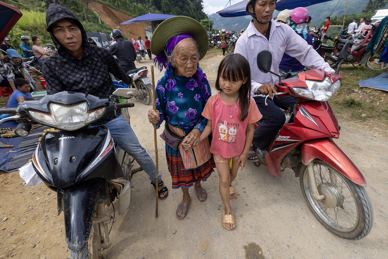 A group of people, including an elderly woman and a young girl holding hands, stand between two motorbikes on a dirt road. The scene is bustling with people in Norhtern Vietnam.