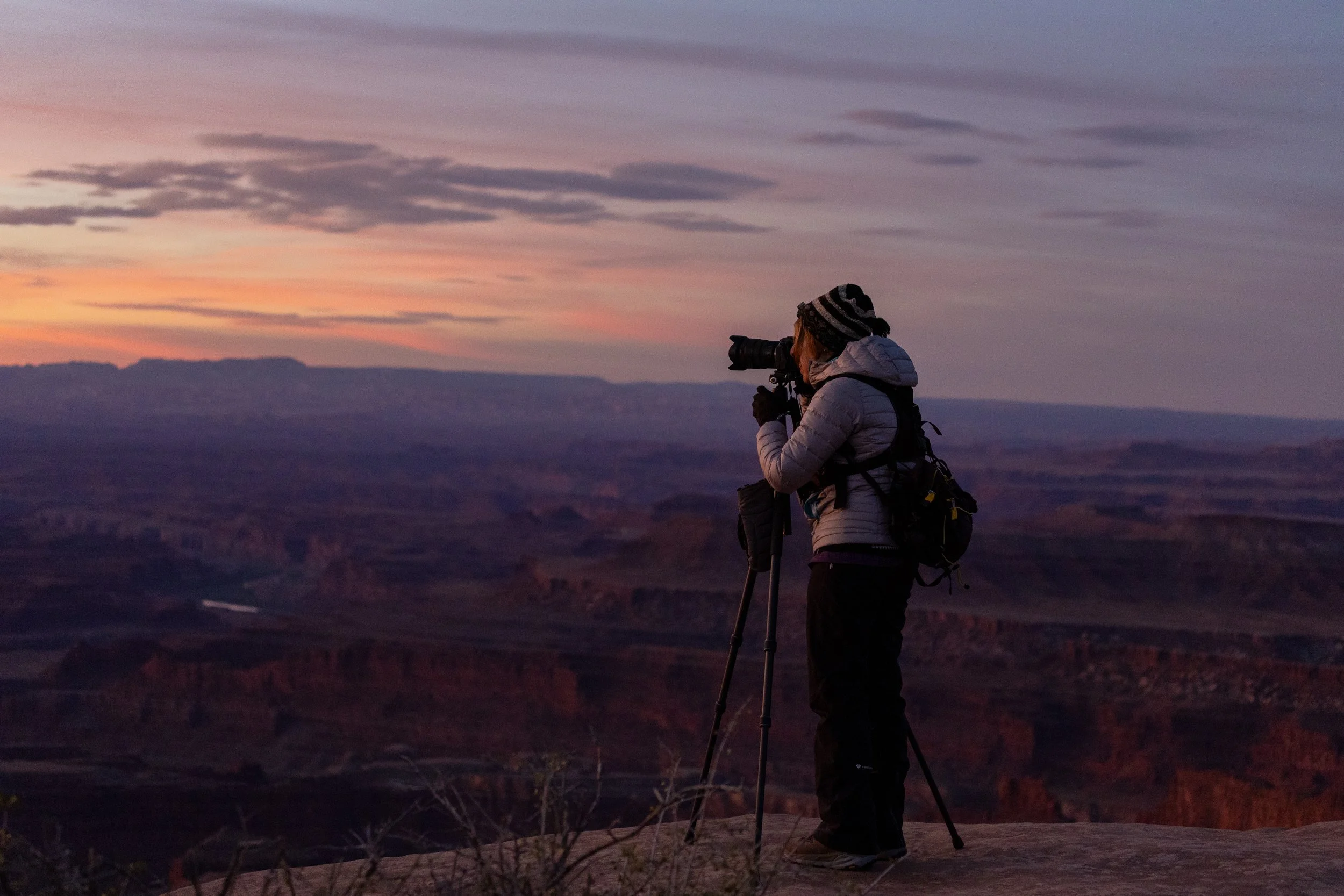 A person with a camera on a tripod standing on a rocky ledge at sunset or sunrise, overlooking a canyon landscape.