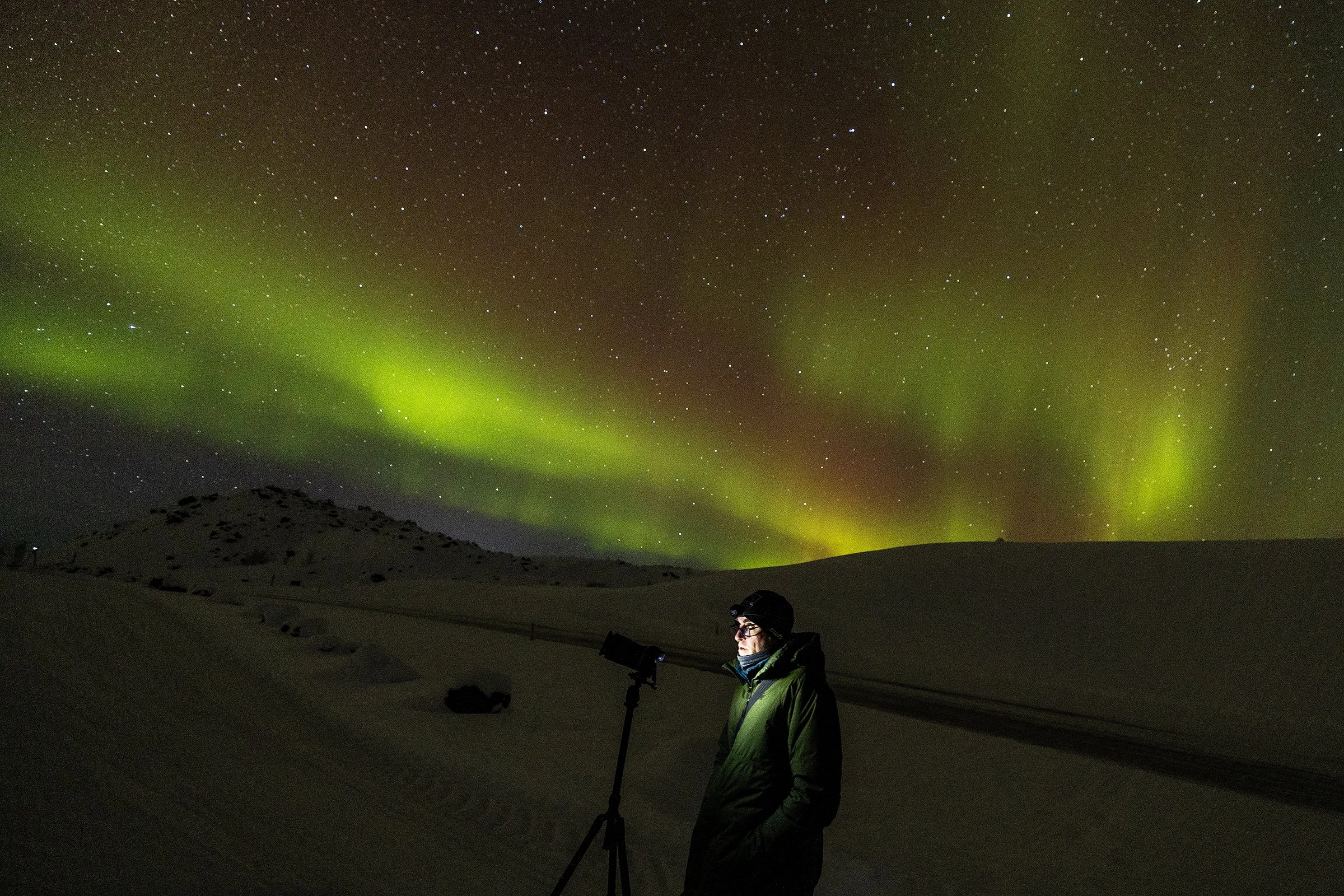 A person in winter clothing standing outdoors at night, looking at the northern lights in a snowy landscape in Iceland.