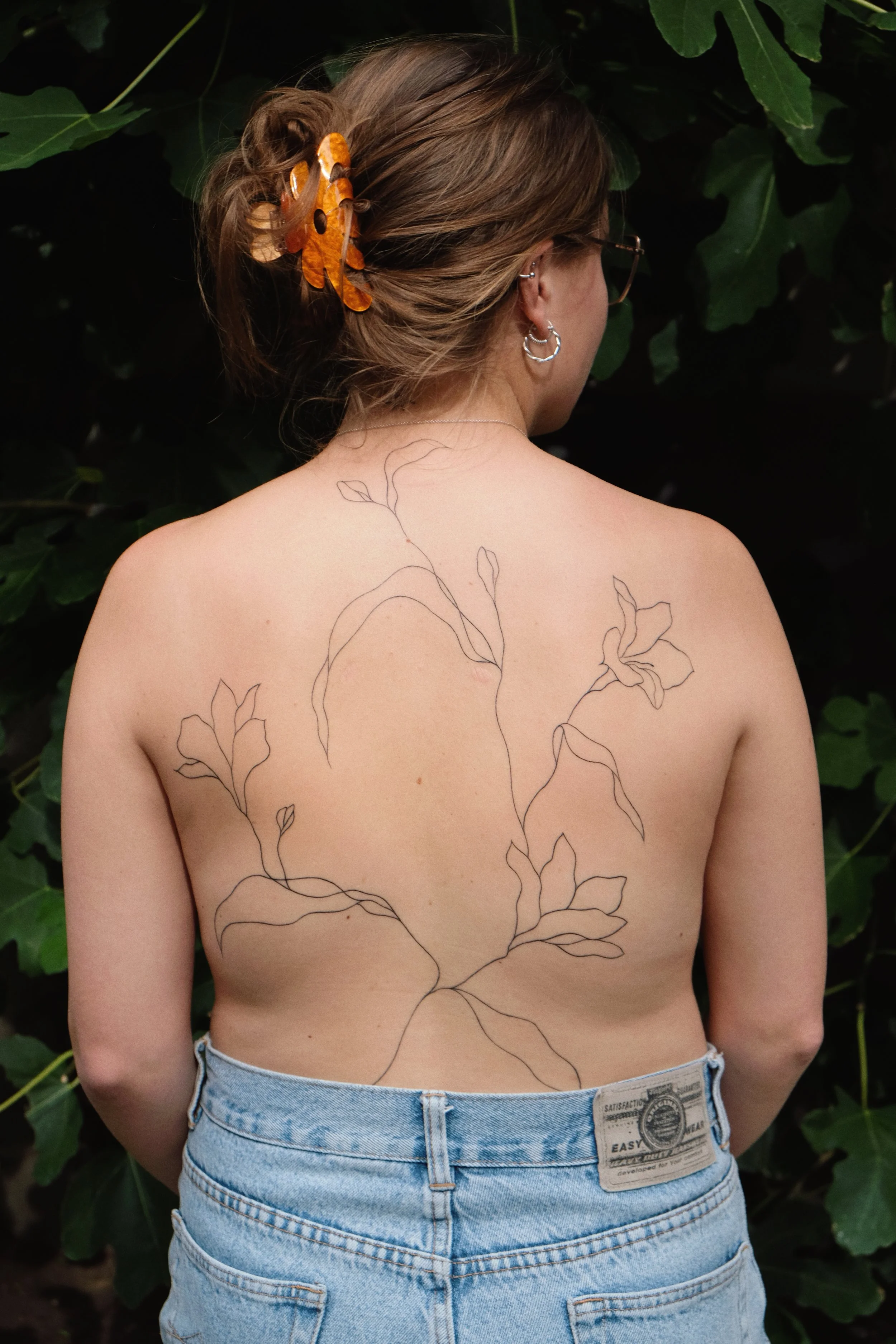 A woman with light brown hair styled in a bun with orange and black butterfly hair clips, wearing glasses and silver jewelry, overlooks a large tattoo of leafy vines on her bare back. She is wearing light blue jeans with a tag visible on the waistban
