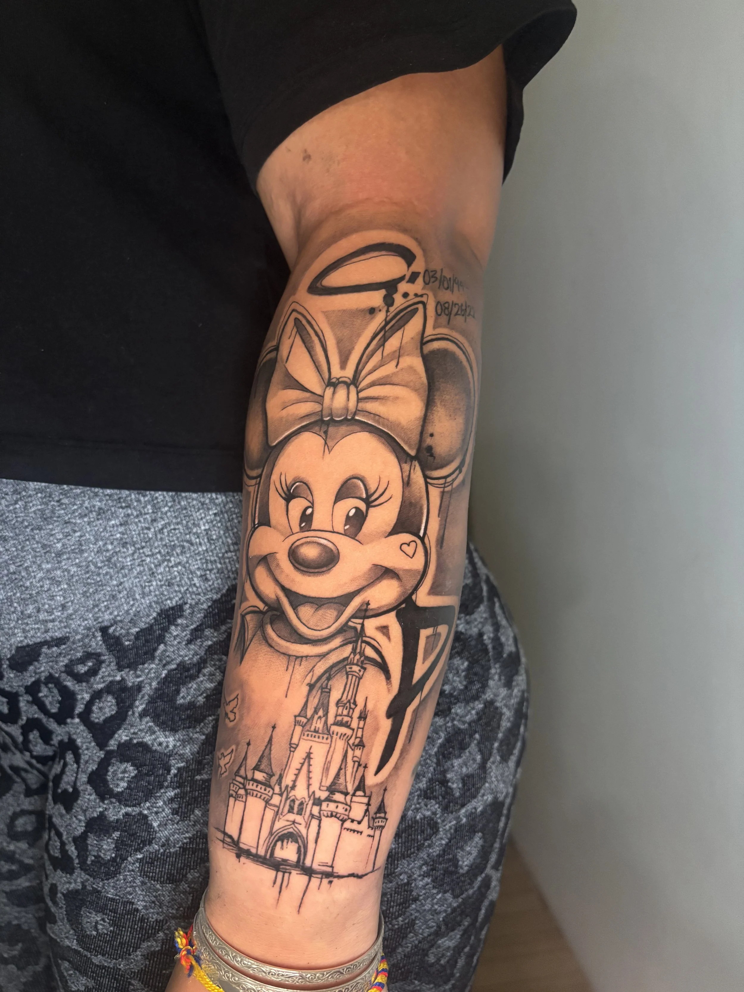 A tattoo of a Disney character, Minnie Mouse, on a person's forearm, with a castle below and surrounding details.