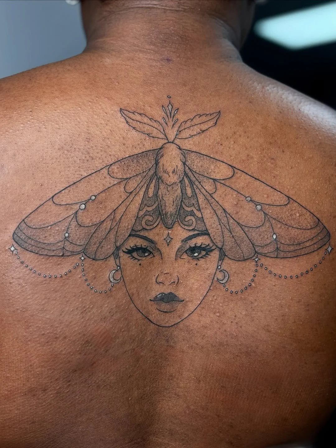 Tattoo of a woman's face with decorative jewelry and a large moth on her head, detailed with wings and antennae, on a person's back.