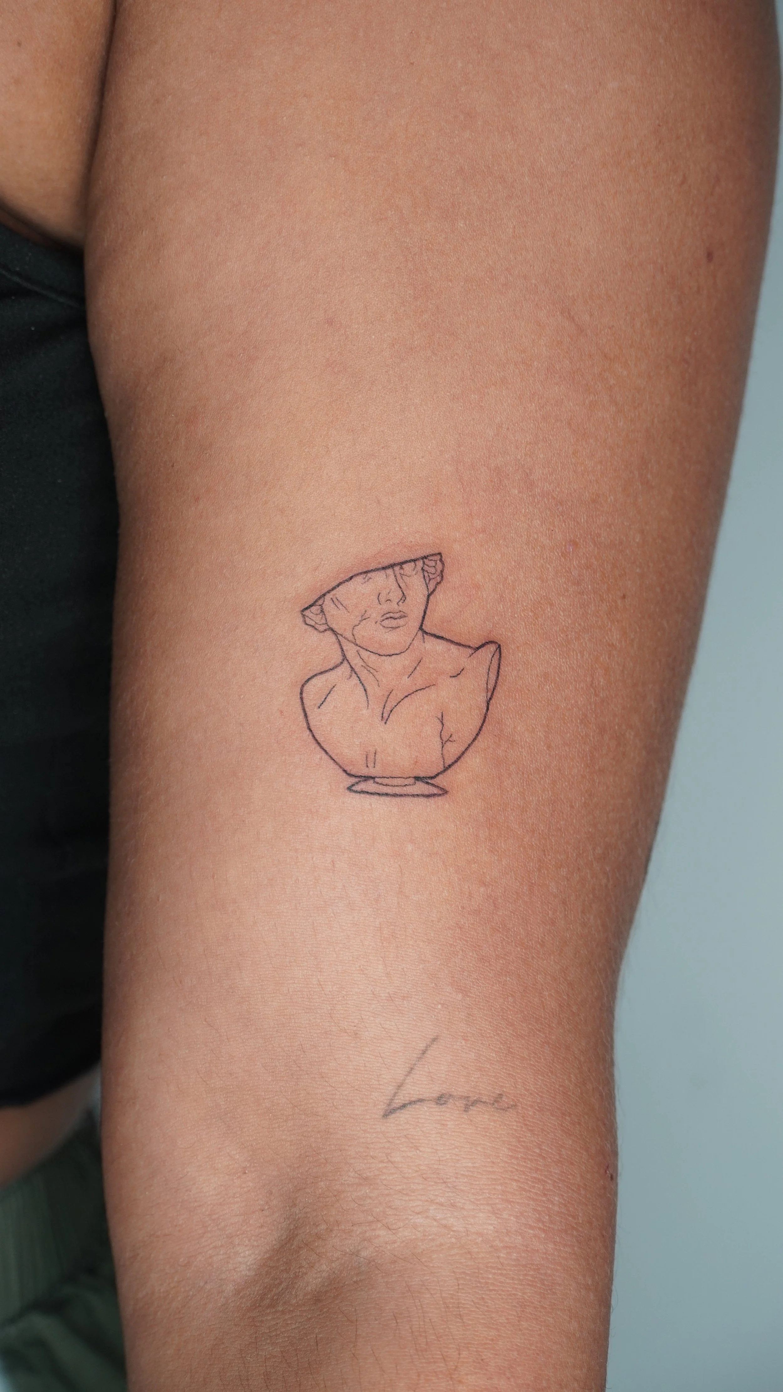 A tattoo of a minimalist line drawing of a classical bust of a woman on a person's upper arm, with the word 'Love' written in cursive below.