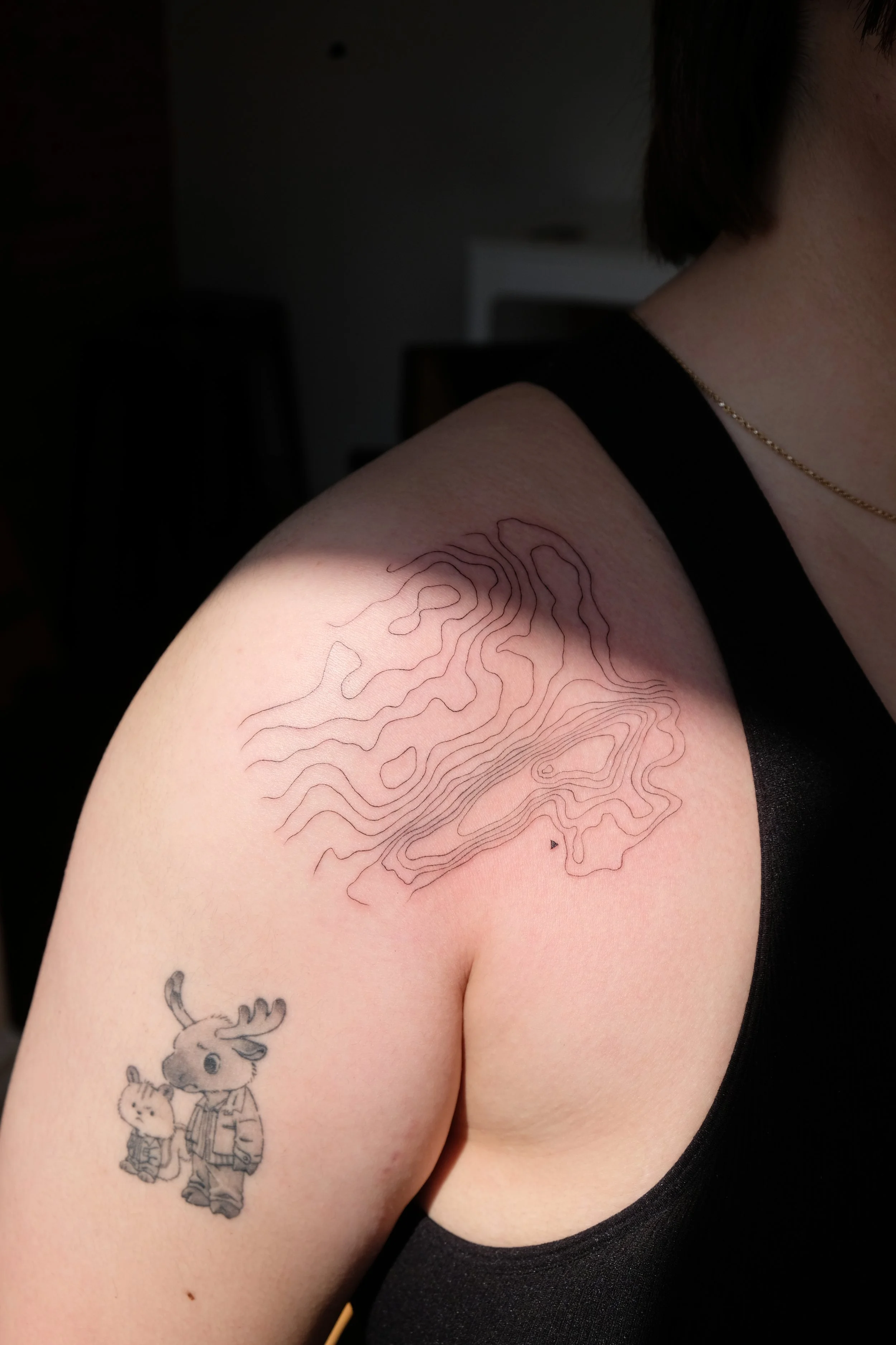 Close-up of a person's shoulder with a topographic line tattoo on the skin, a cartoon-style tattoo of two animals, and part of their face and black clothing visible.