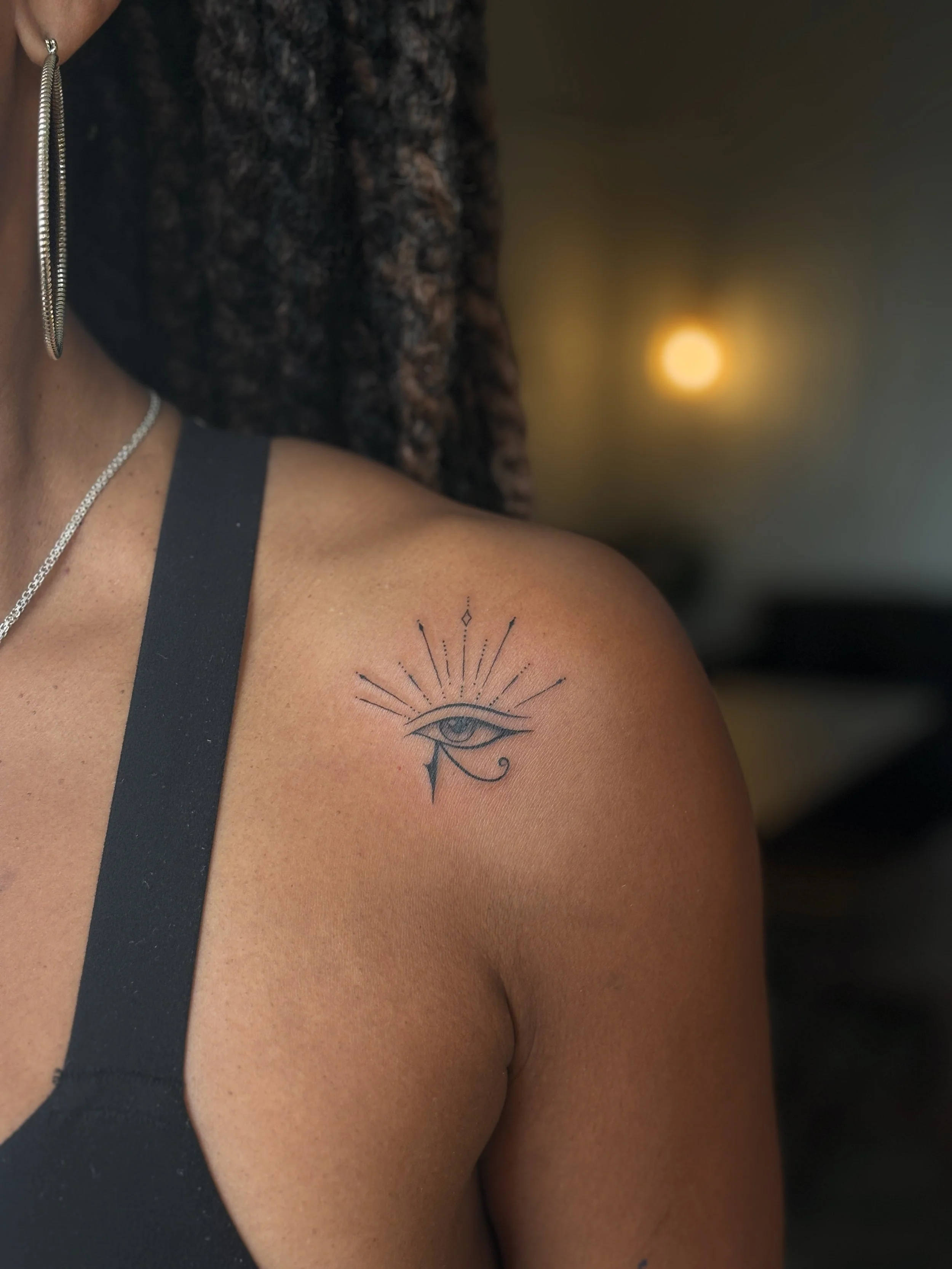 A close-up of a person's shoulder showing a black tattoo of an eye with rays extending outward, with jewelry visible on the person's neck and ear, in a dimly lit environment with warm lighting.