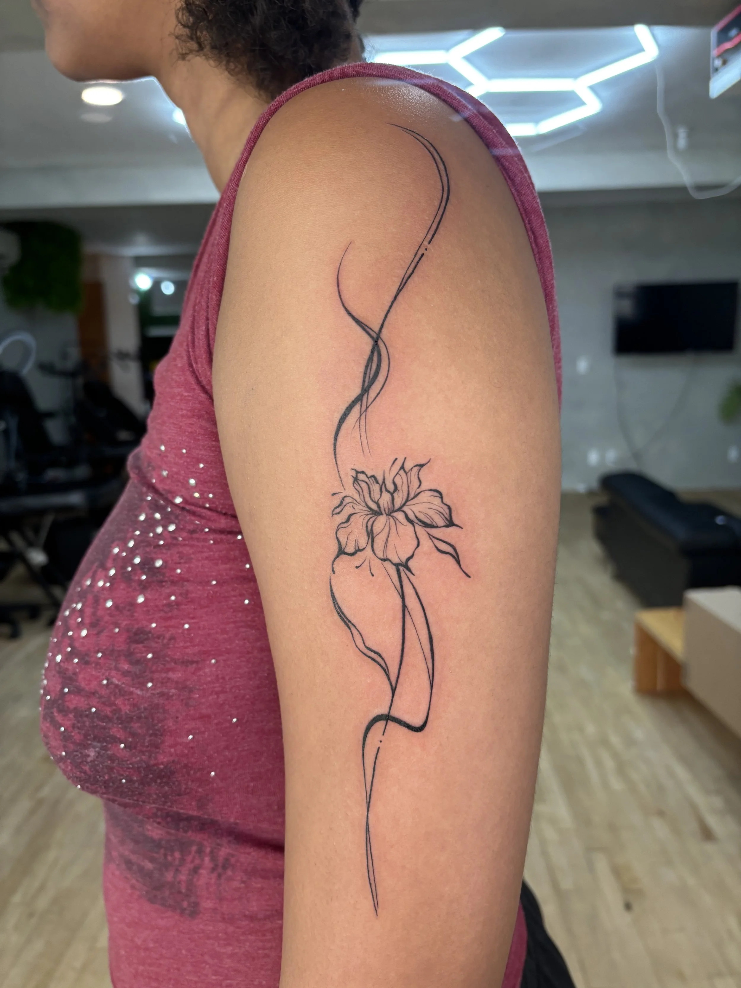 A person's upper arm with a black floral tattoo and swirling line design, wearing a maroon sleeveless top with small rhinestones.