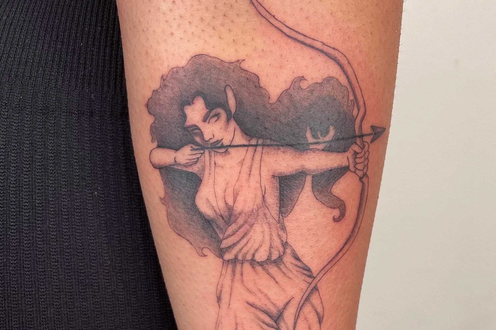 Tattoo of Cupid aiming an arrow with a bow, with a woman in a flowing dress and curly hair.