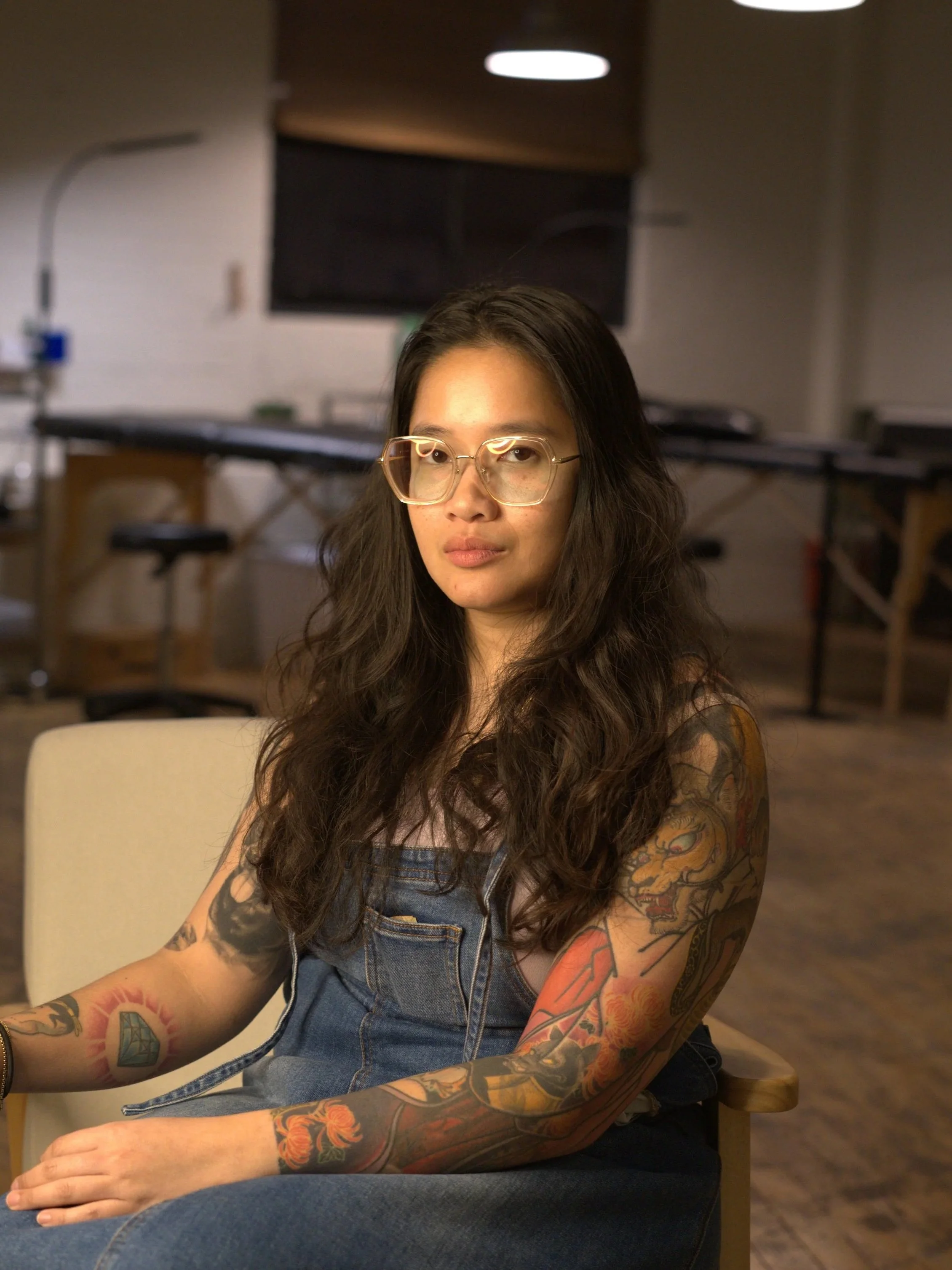 A young woman with long dark wavy hair wearing glasses and a denim overall sits at a table in a room with a kitchen in the background.