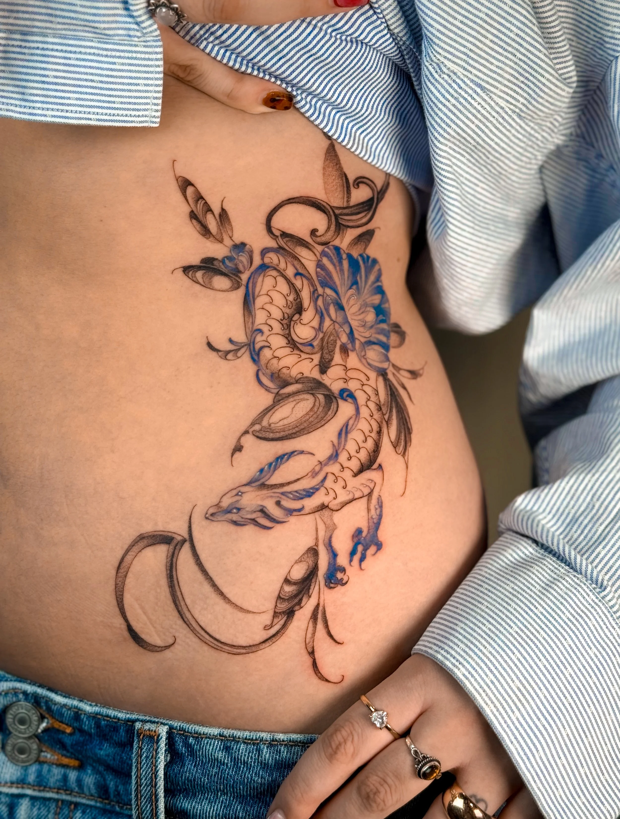 Close-up of a person's side showing a detailed tattoo of a dragon with blue accents, partially covered by a striped shirt and jewelry including rings and a bracelet.