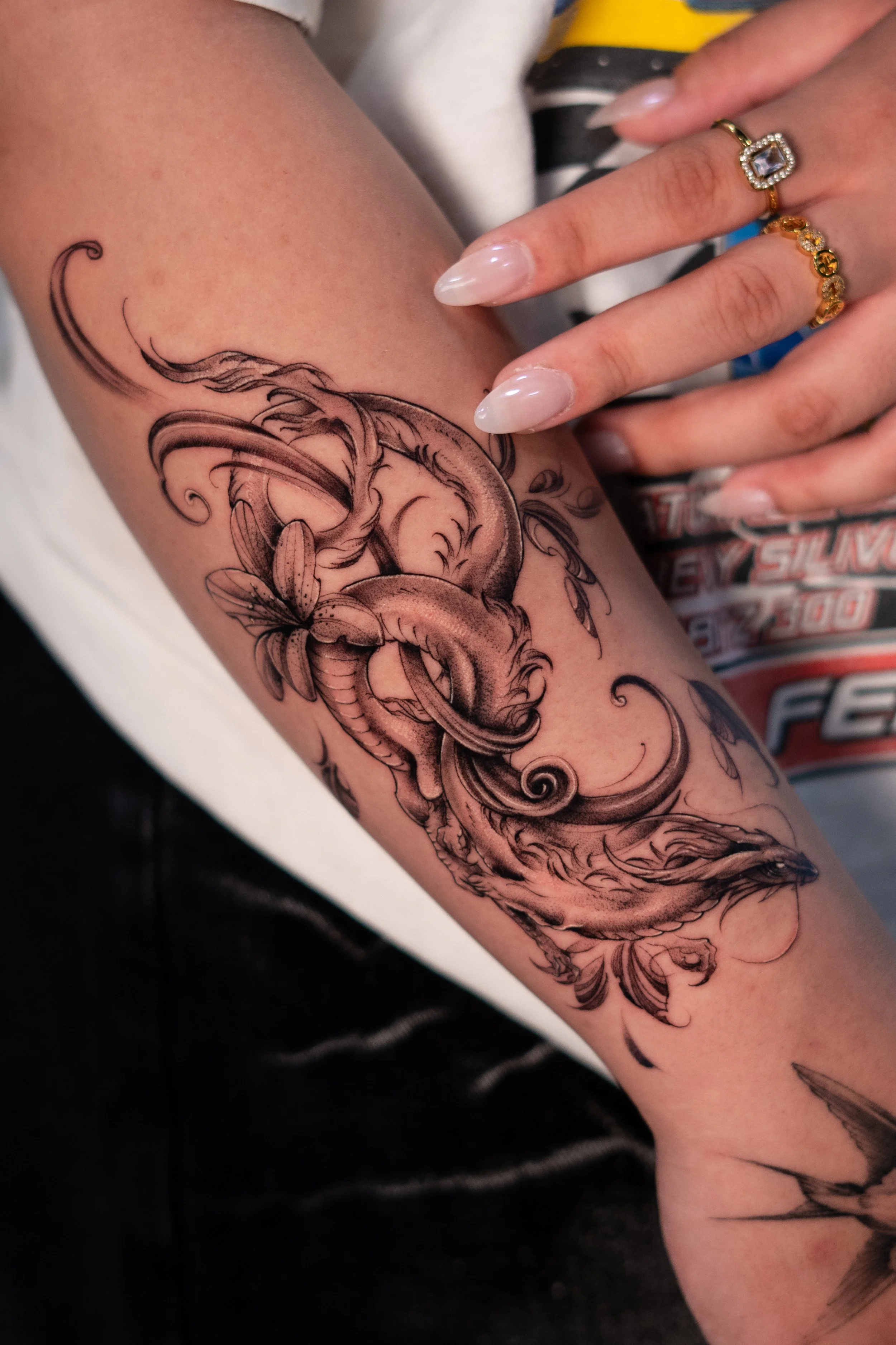 Close-up image of a person's arm with a detailed black and white tattoo of a whale, a flower, and flowing waves. The person's hand, adorned with two gold rings with decorative stones, rests on the arm. The background shows a patterned shirt and part 