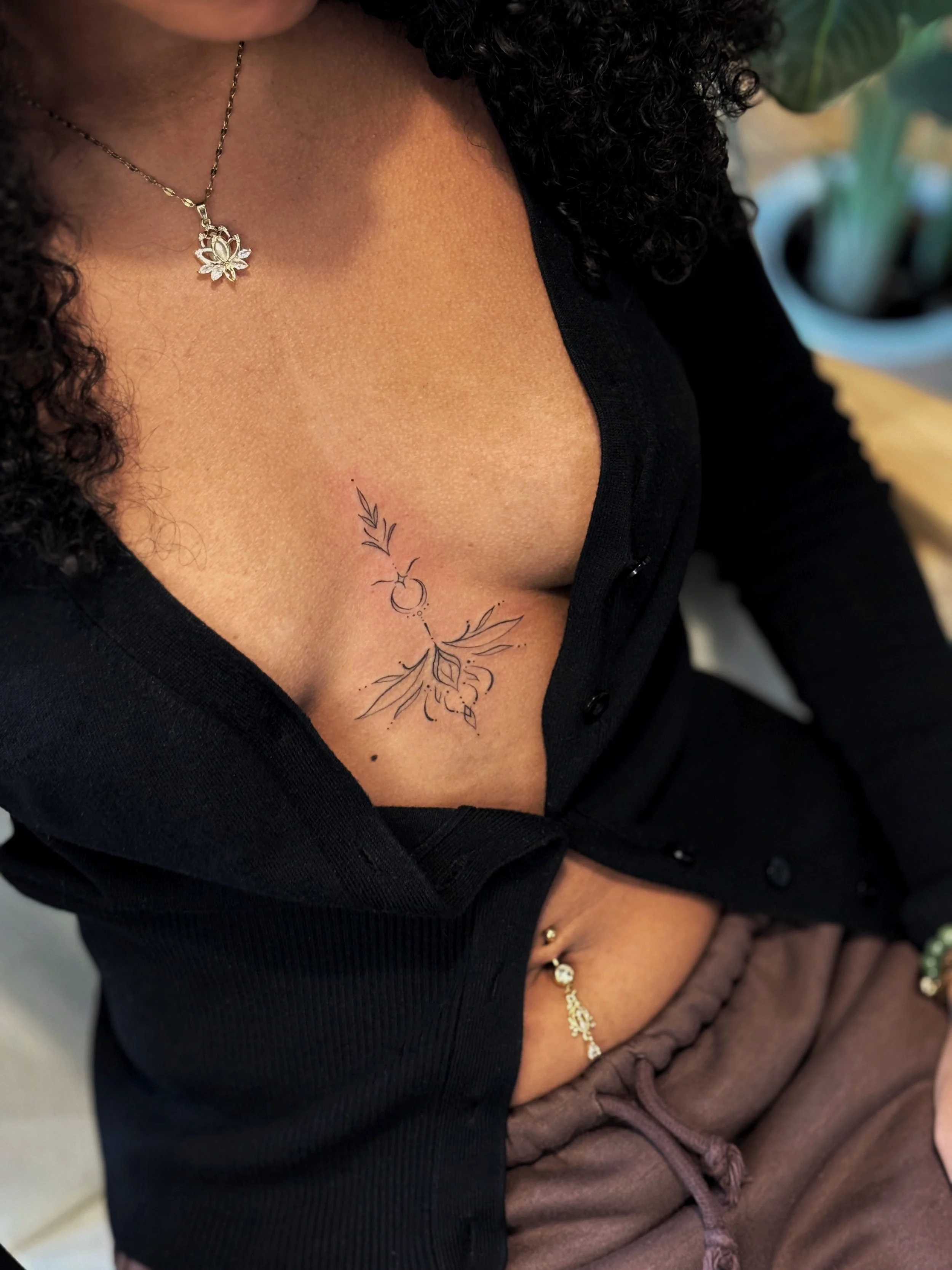 Close-up of a woman's chest showing a tattoo of a bouquet with leaves, a lotus flower, and decorative elements, paired with a silver lotus-shaped necklace, wearing a black top.