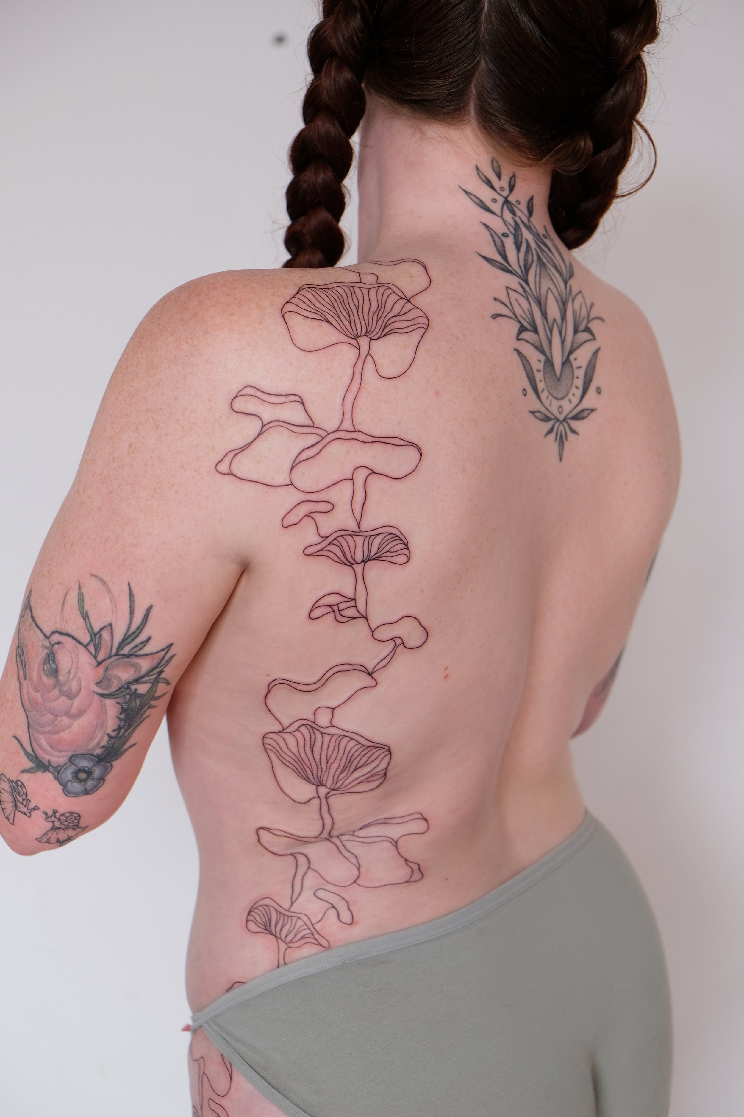 A woman with braided hair showing her back and shoulder tattoos, including line art mushrooms on her shoulder and floral designs on her back.