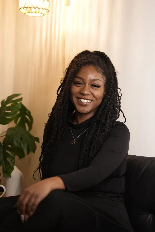 Smiling woman with long curly hair sitting on a black leather chair in a cozy room, next to a green potted plant and beige curtains.