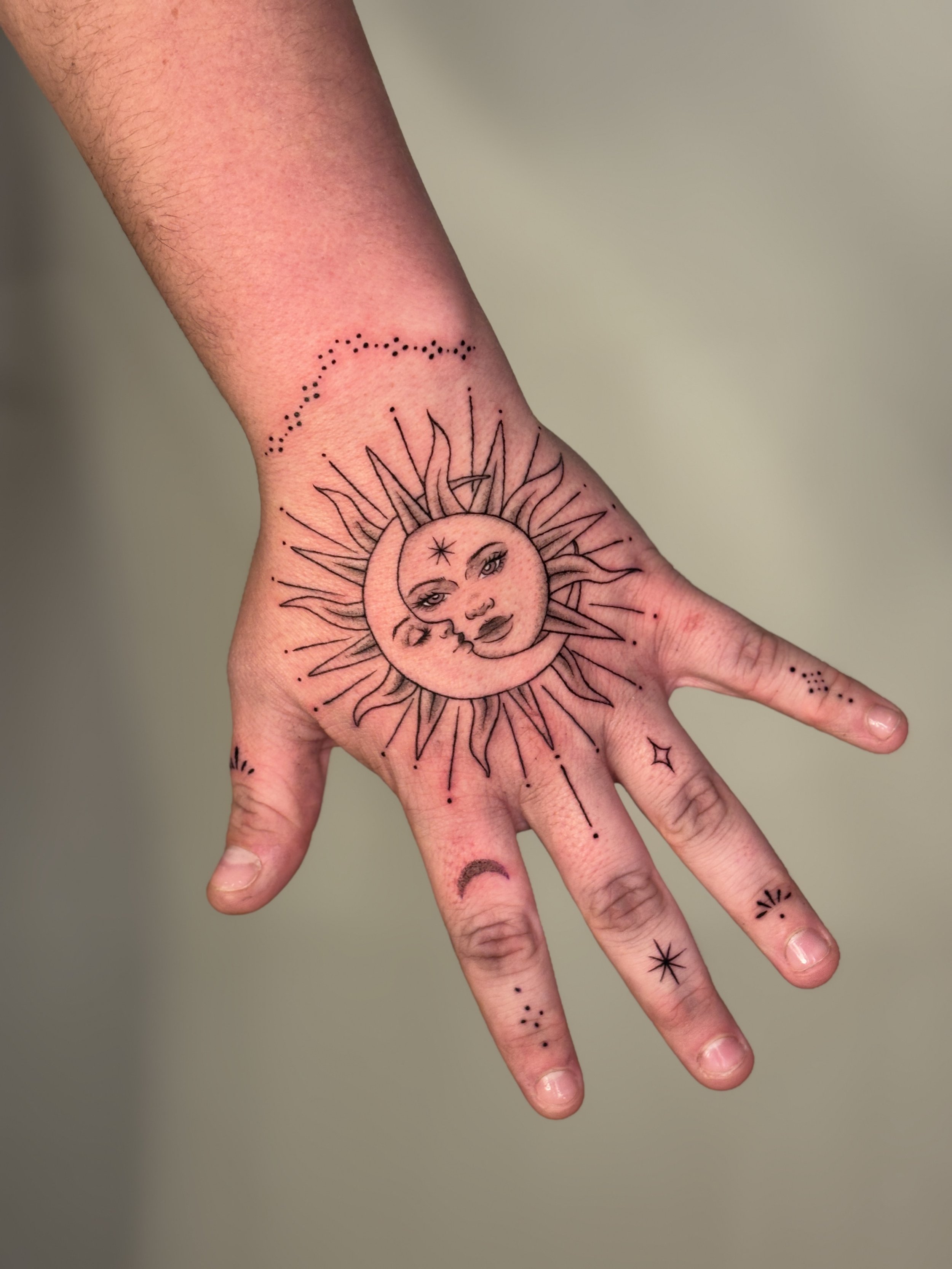 Tattoo of the sun with a face, moon, stars, and solar system symbols on a person's hand and fingers.