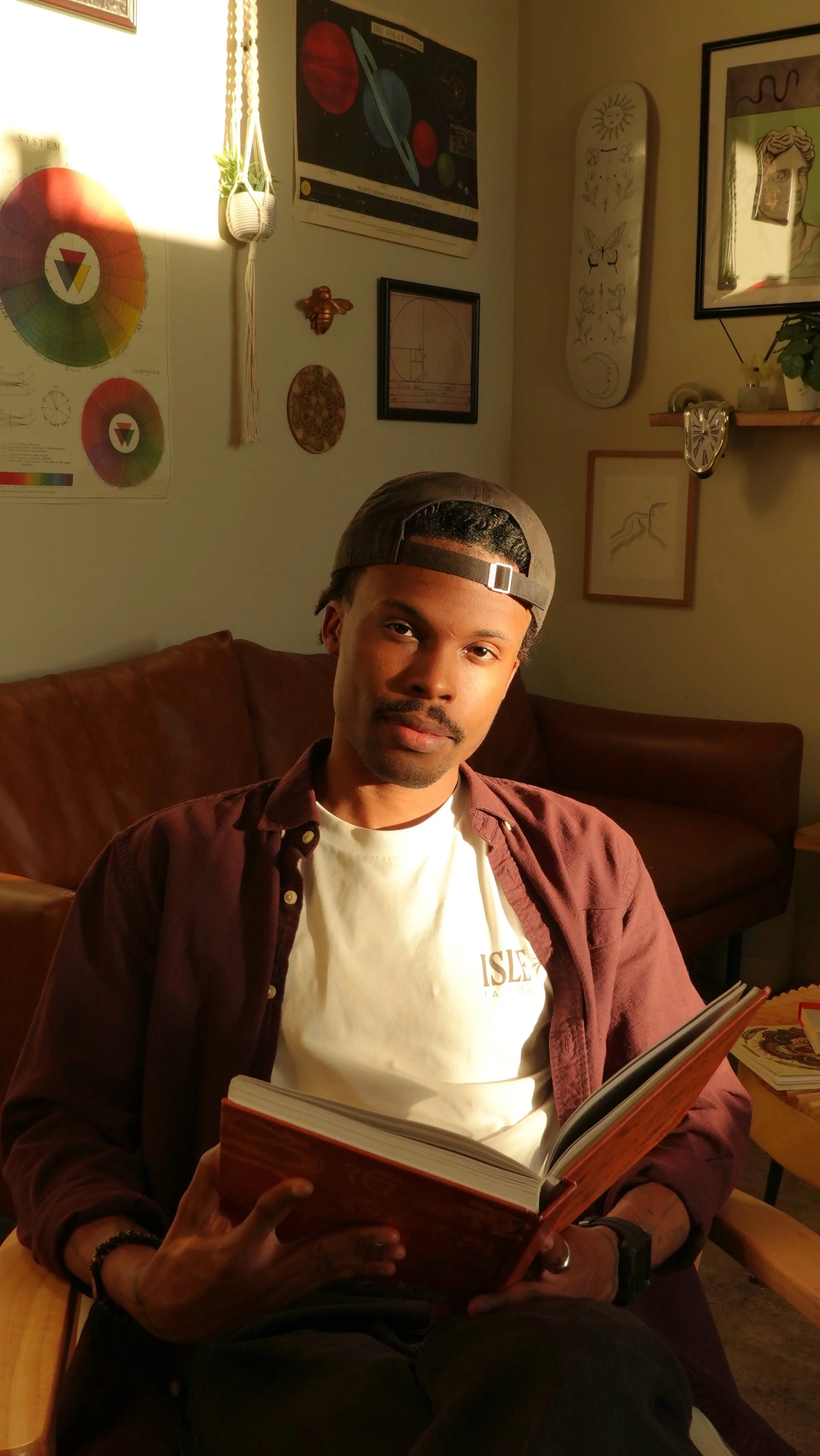 Young man in a backward baseball cap reading an open book on a couch in a warmly lit room with colorful artwork and plants.