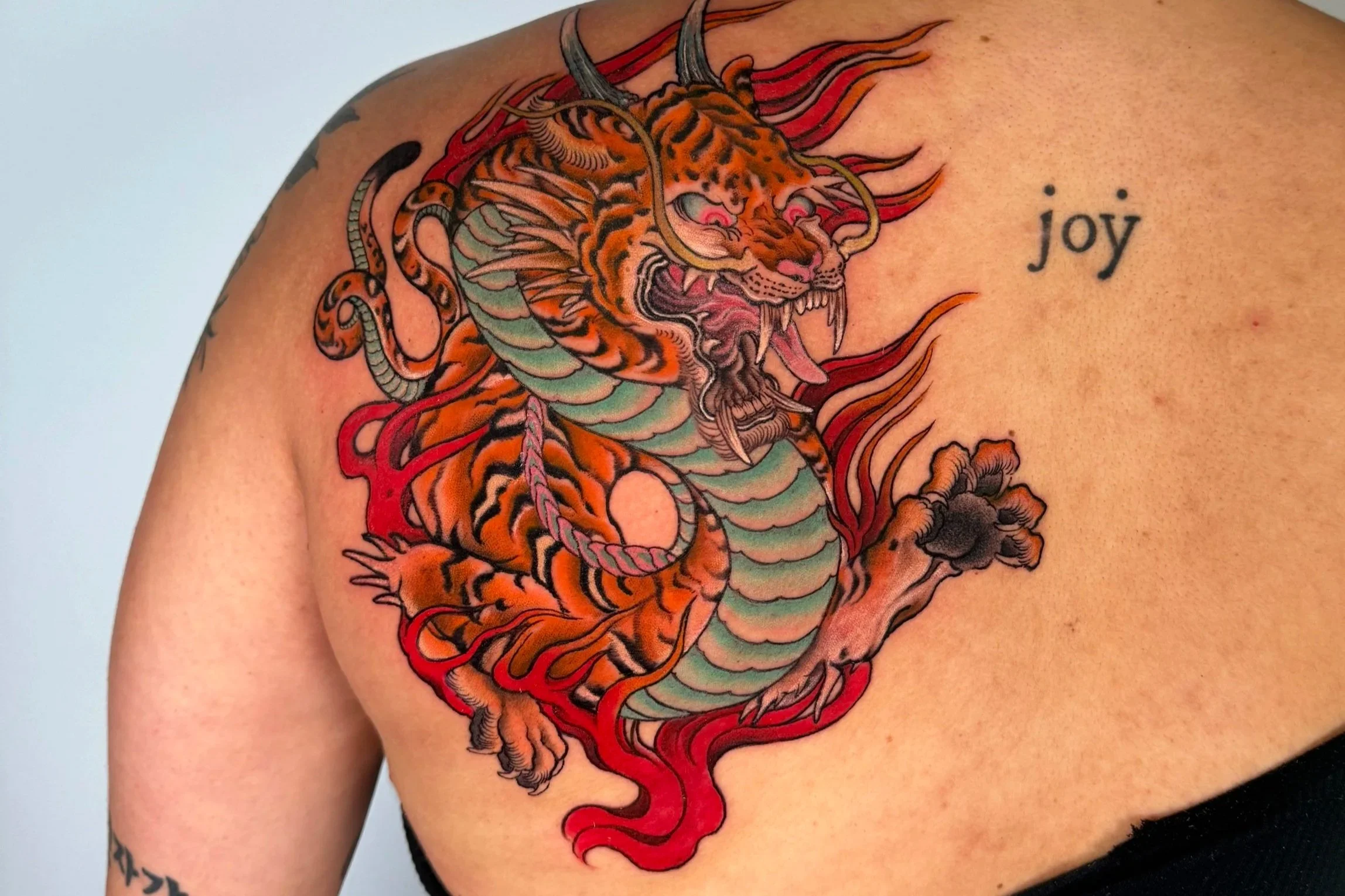 Tattoo of a fierce tiger with a green body and orange stripes, surrounded by red flames, on a person's shoulder. The word 'joy' is tattooed nearby.
