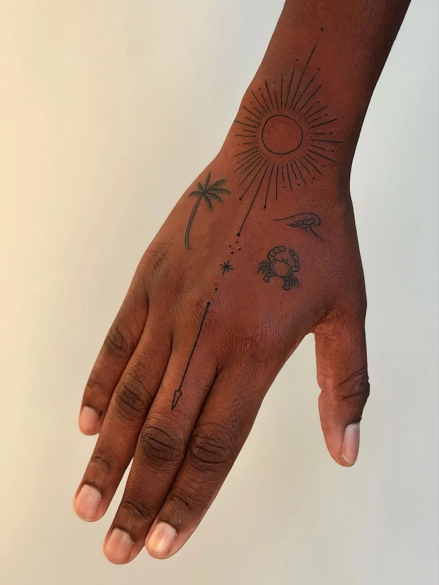 Close-up of a hand with minimalist nature-themed tattoos, including a sun, palm tree, crocodile, crab, star, and arrow.