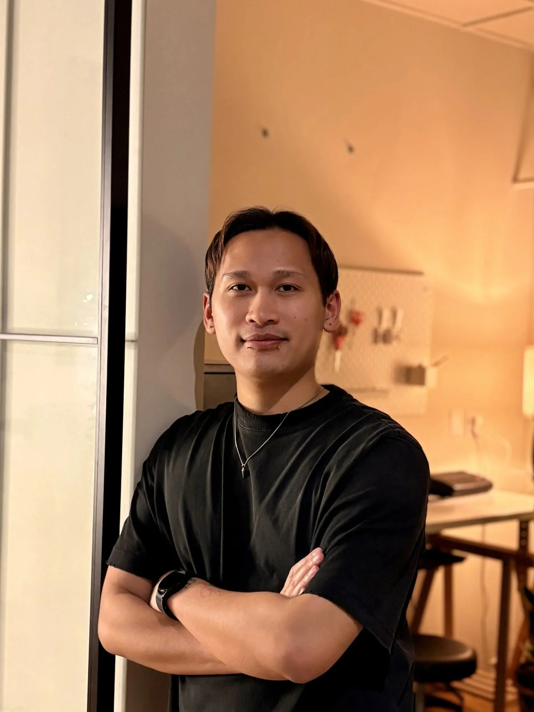 A young man with crossed arms, standing indoors in front of a wall and a mirror, wearing a black t-shirt and a necklace, smiling slightly.