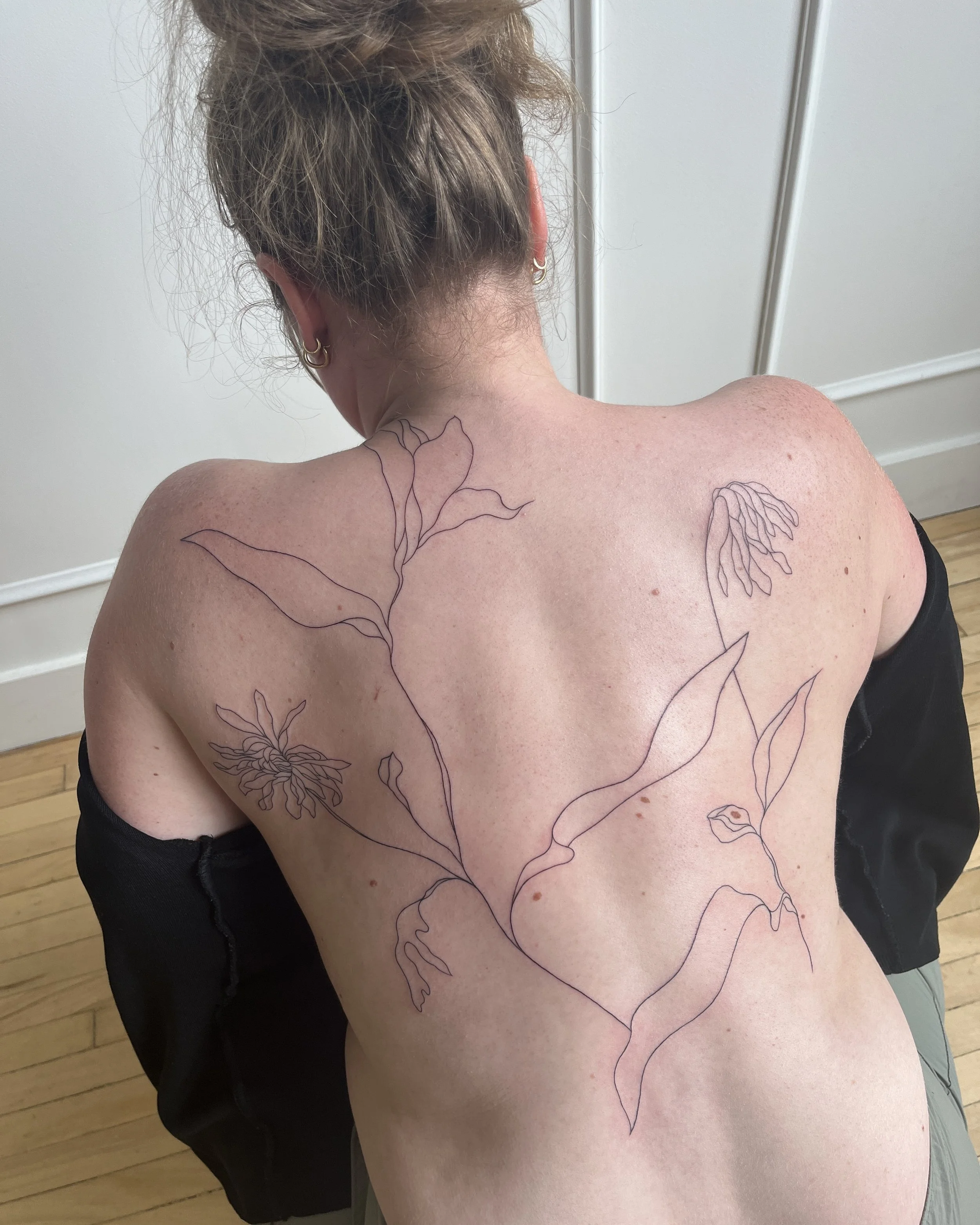 A woman with shoulder-length hair tied up in a bun, wearing earrings, is showing her back and shoulder with a large floral tattoo outline drawn with black ink on her skin.