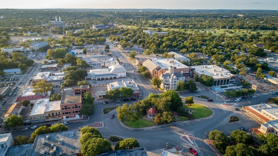 Aerial view of a small town with a central park surrounded by historic buildings, retail stores, and parking lots, with tree-lined streets and green spaces.