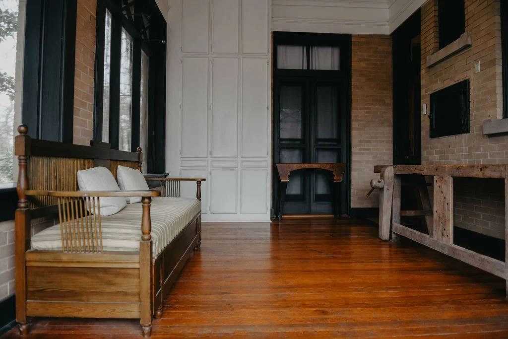 Interior of a room with wooden furniture, a bench with striped cushions and two pillows, large windows, brick walls, and a wooden workbench.
